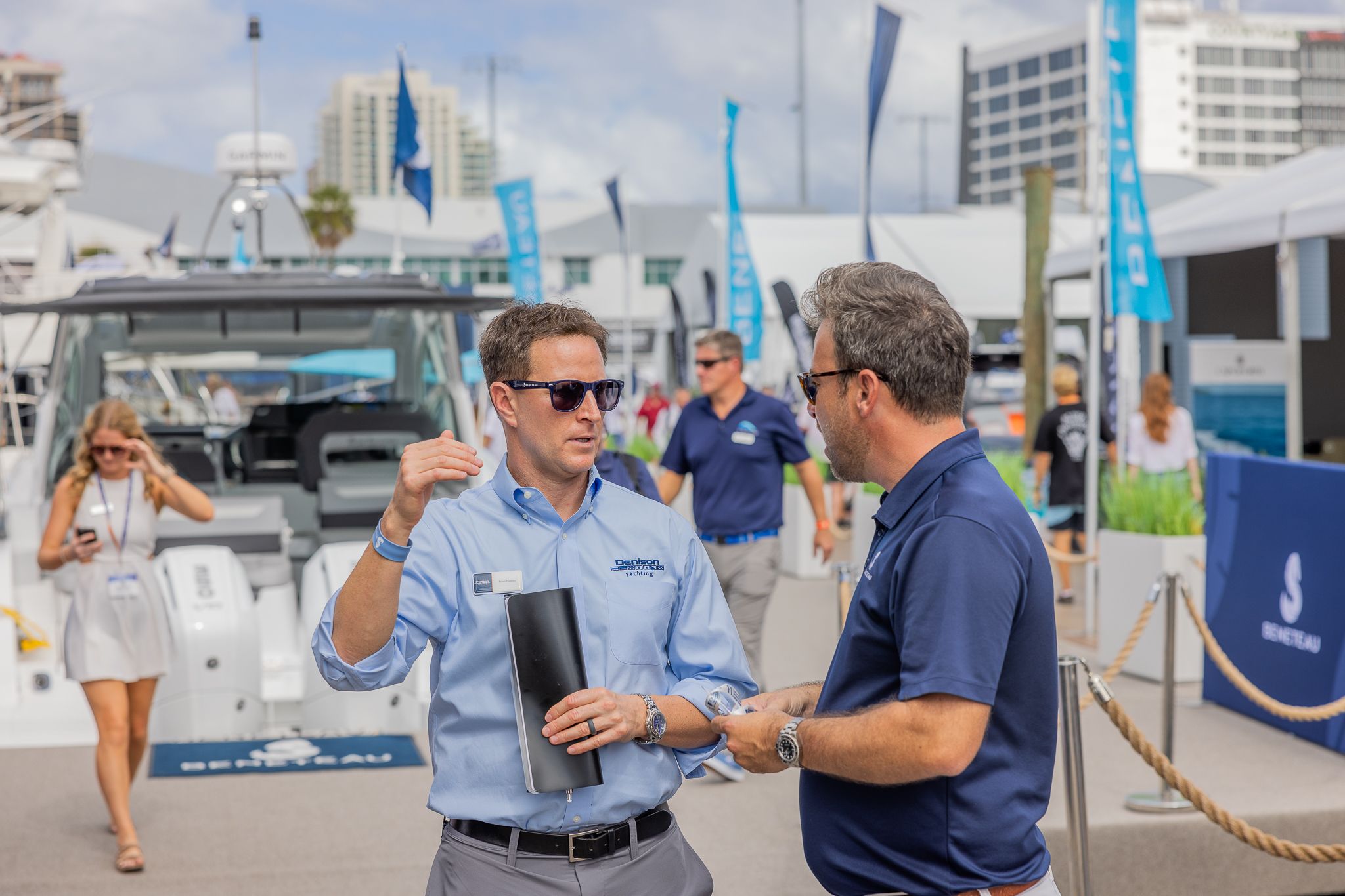 Two men conversing at an outdoor boat show with boats and buildings in the background.