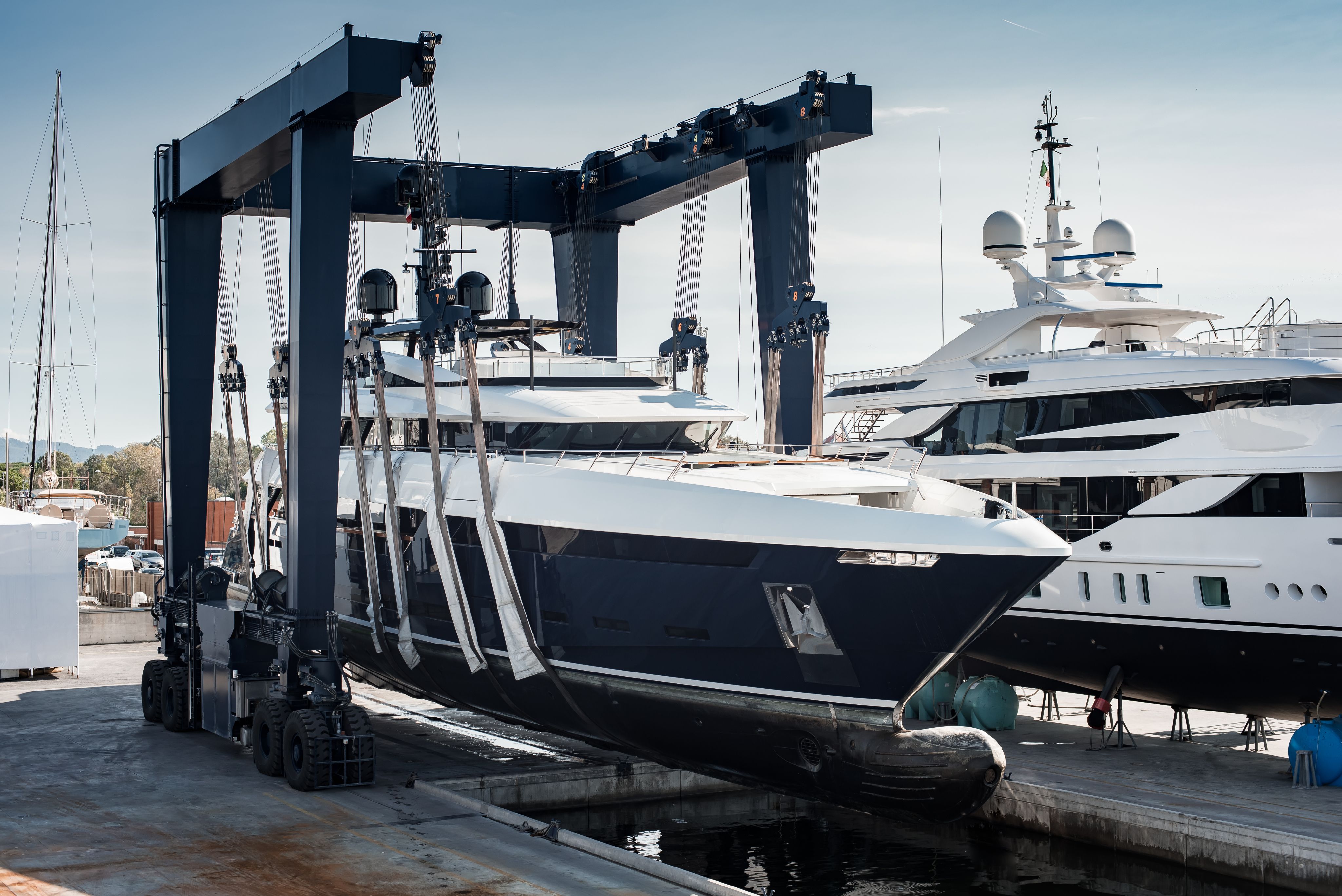 Large blue and white yacht being lifted by a gantry crane in a bustling shipyard.