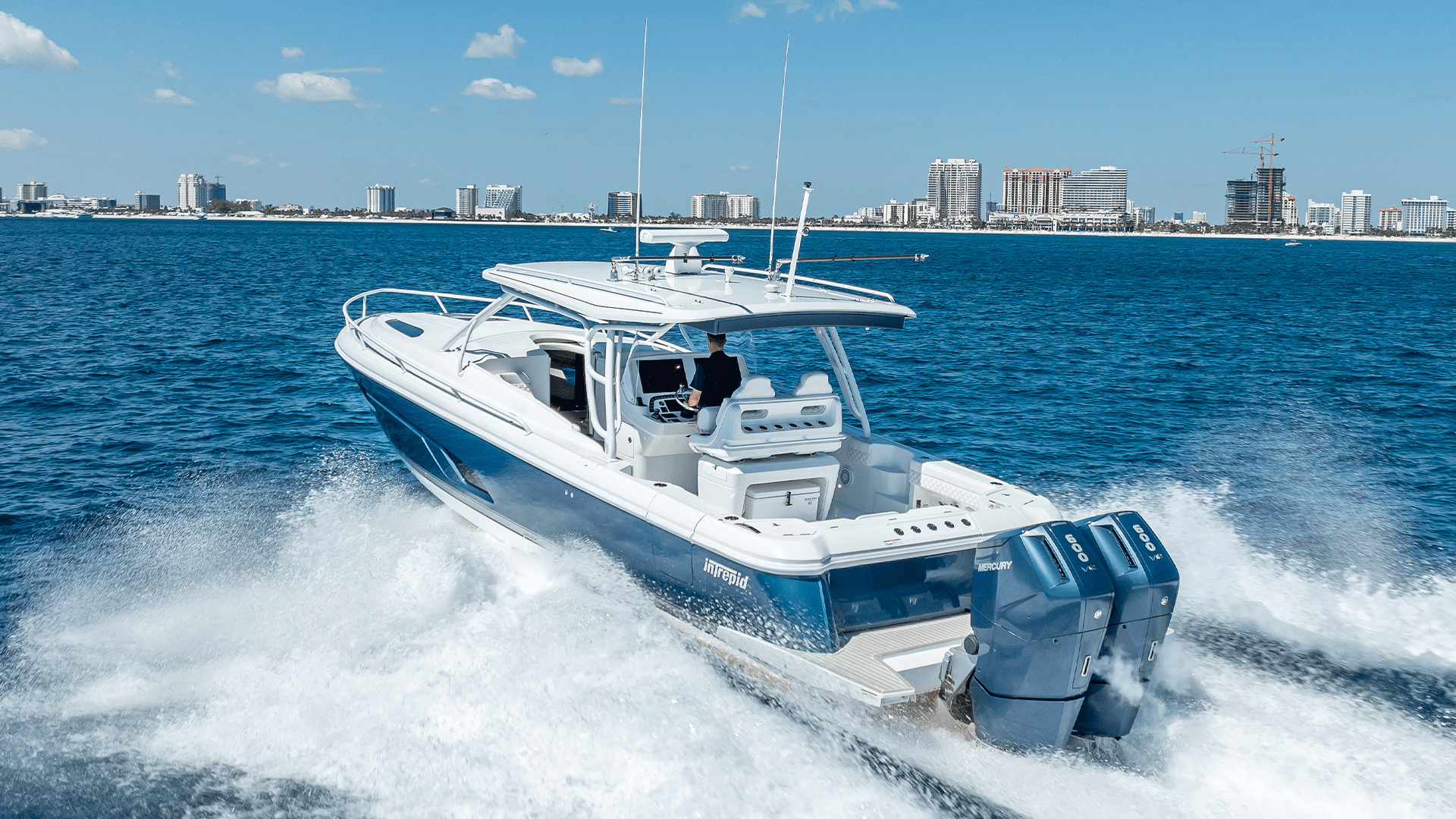 White and blue powerboat speeding across water with a city skyline in the background.