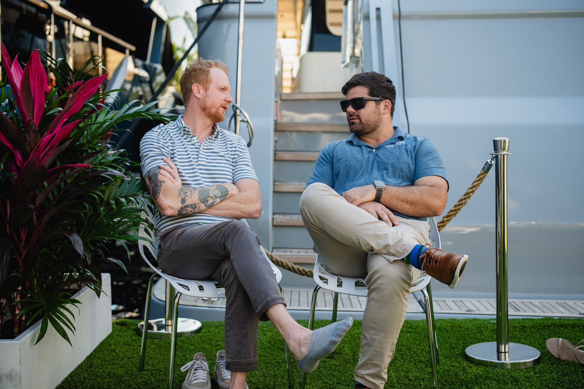 Two men sitting on chairs outdoors, talking on artificial grass next to a boat.