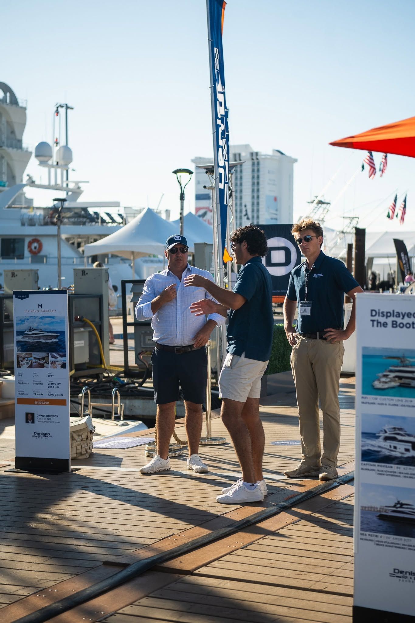 Three men discuss products at an outdoor marine industry event on a sunny dock.