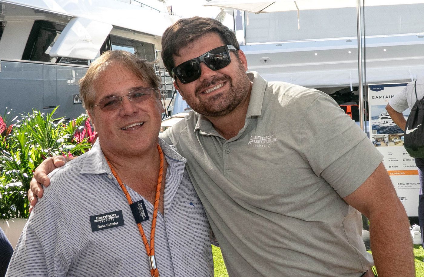 Two smiling men, one wearing sunglasses, pose arm-in-arm outdoors at an event.