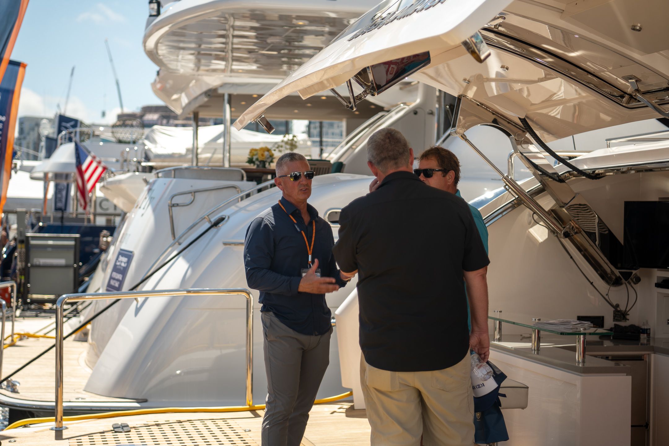 Three men converse on the spacious deck of a large white yacht at a boat show.