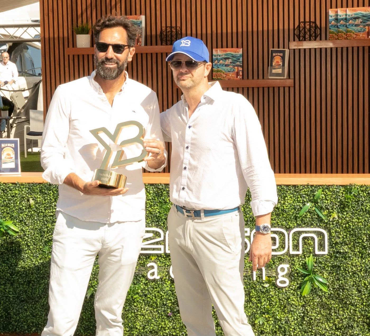 Two men smiling, one holding a trophy, at an outdoor event with a green backdrop.