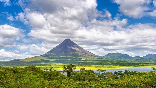 Majestic conical volcano overlooks a tranquil lake and lush tropical forest under a blue sky.