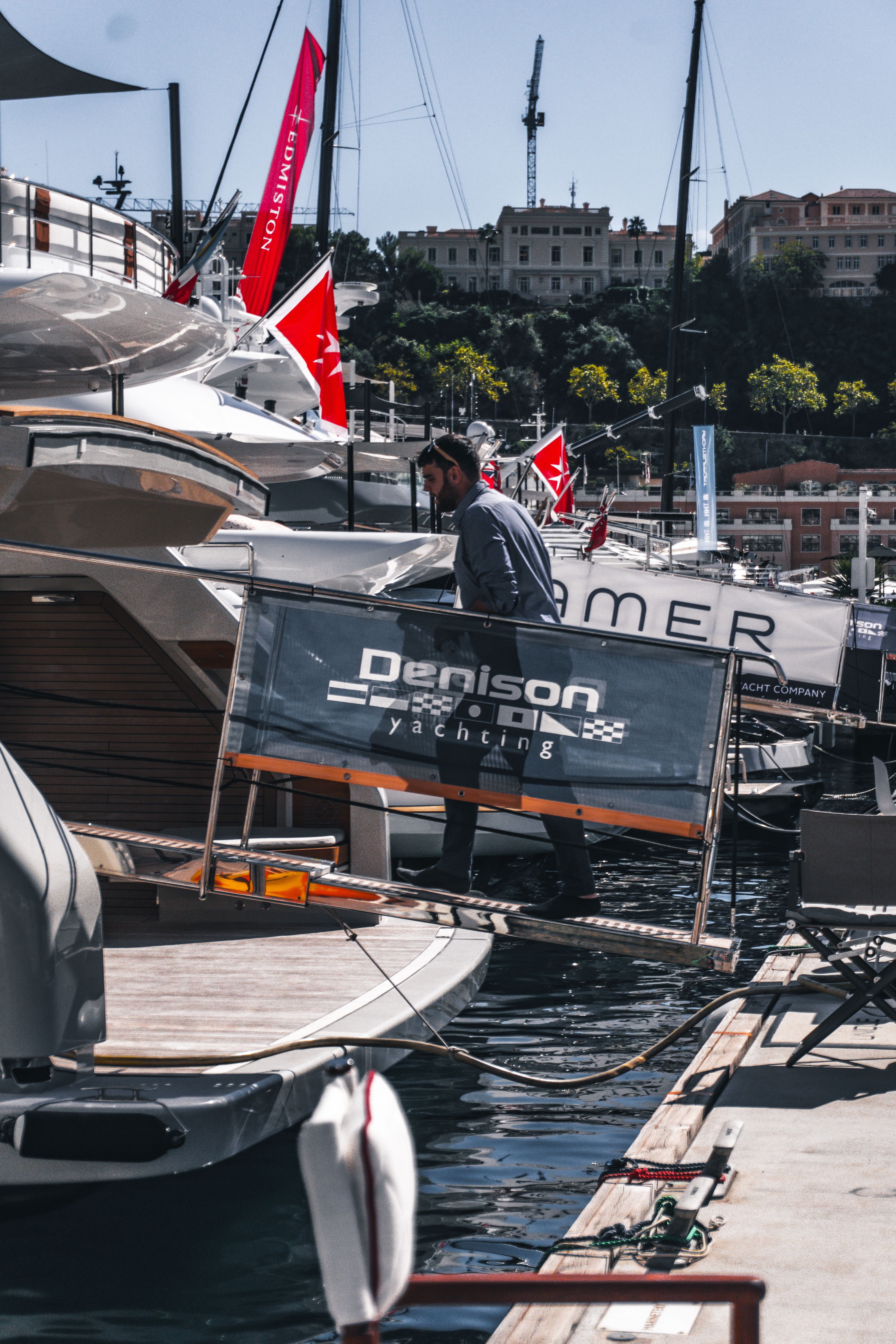 Man sits on boat with 'DENISON' banner in a sunny marina, yachts in background.