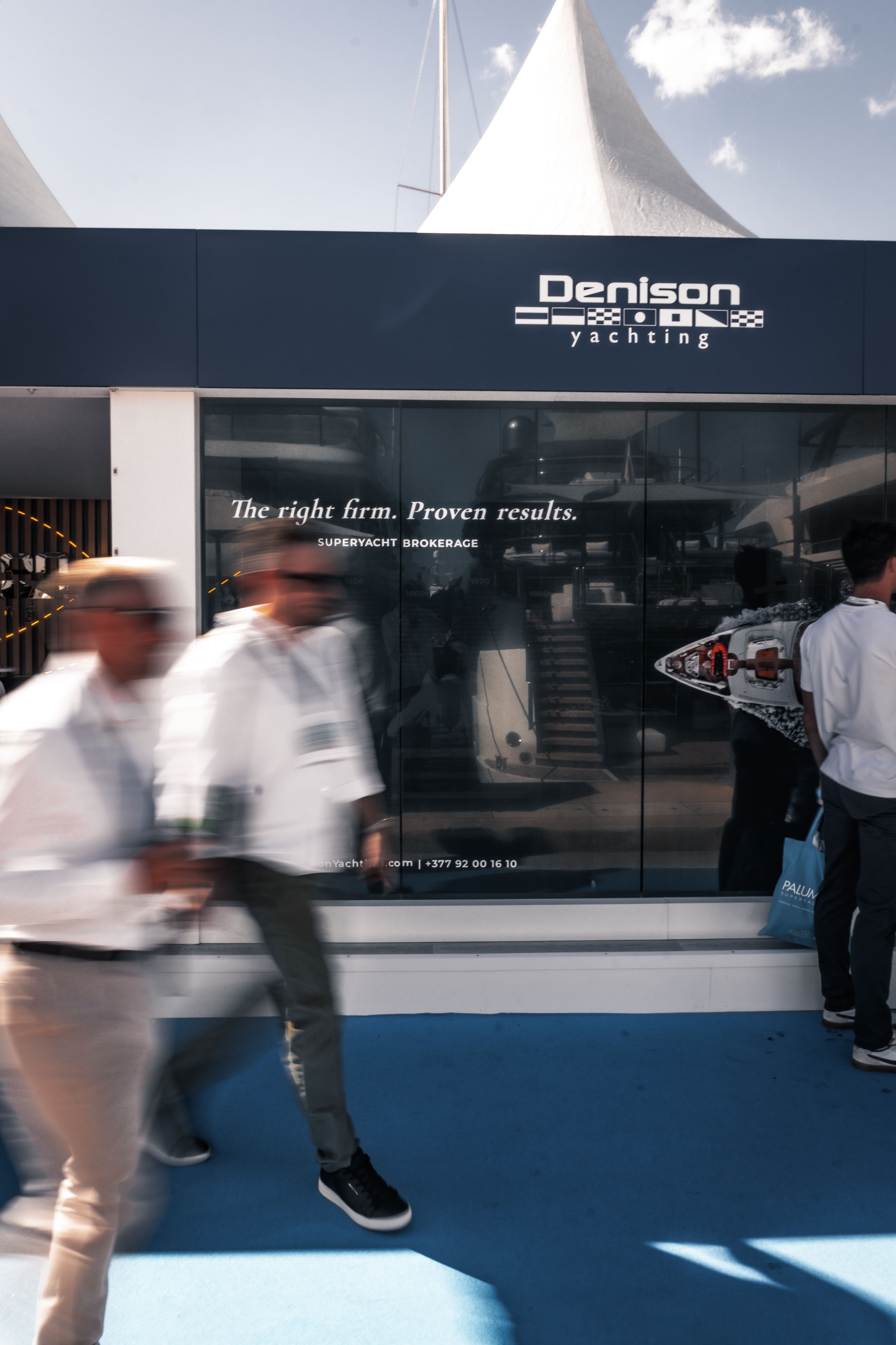 Denison / BERG yacht brokerage booth with blurred people walking by at an outdoor event.