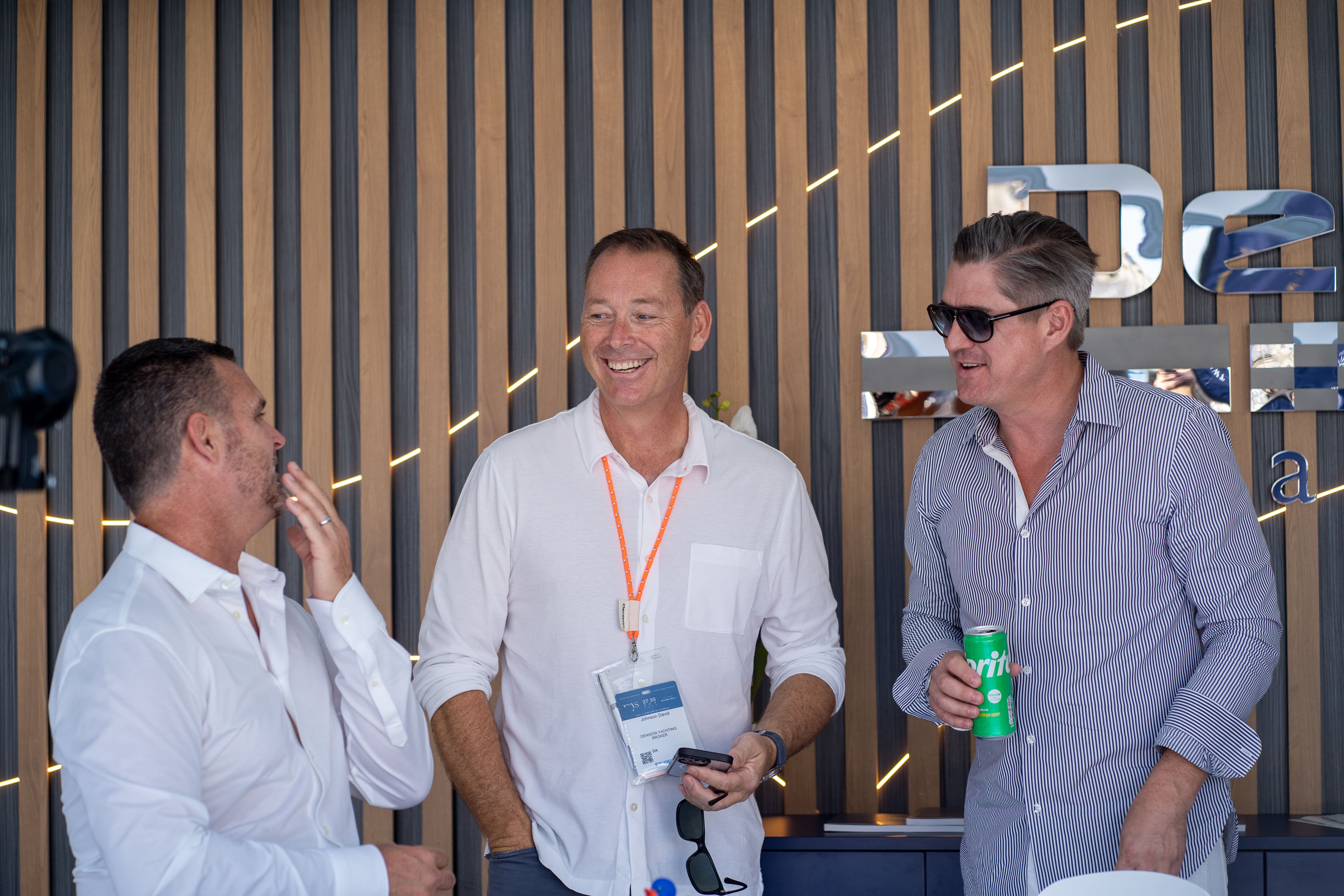 Three men laugh and converse at a social event, one holding a green can.