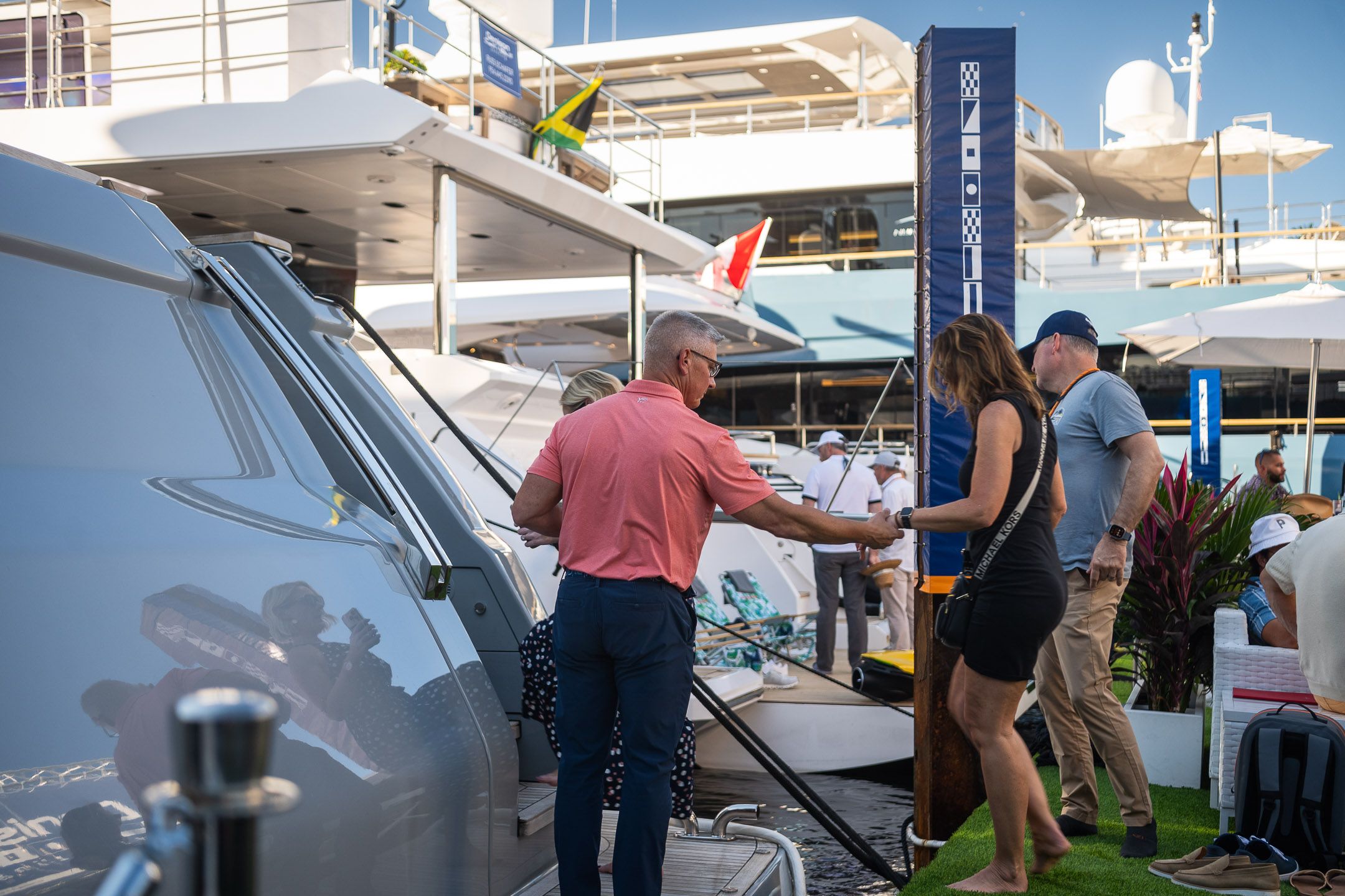 Man assists woman boarding a large silver yacht at an outdoor marine exhibition.
