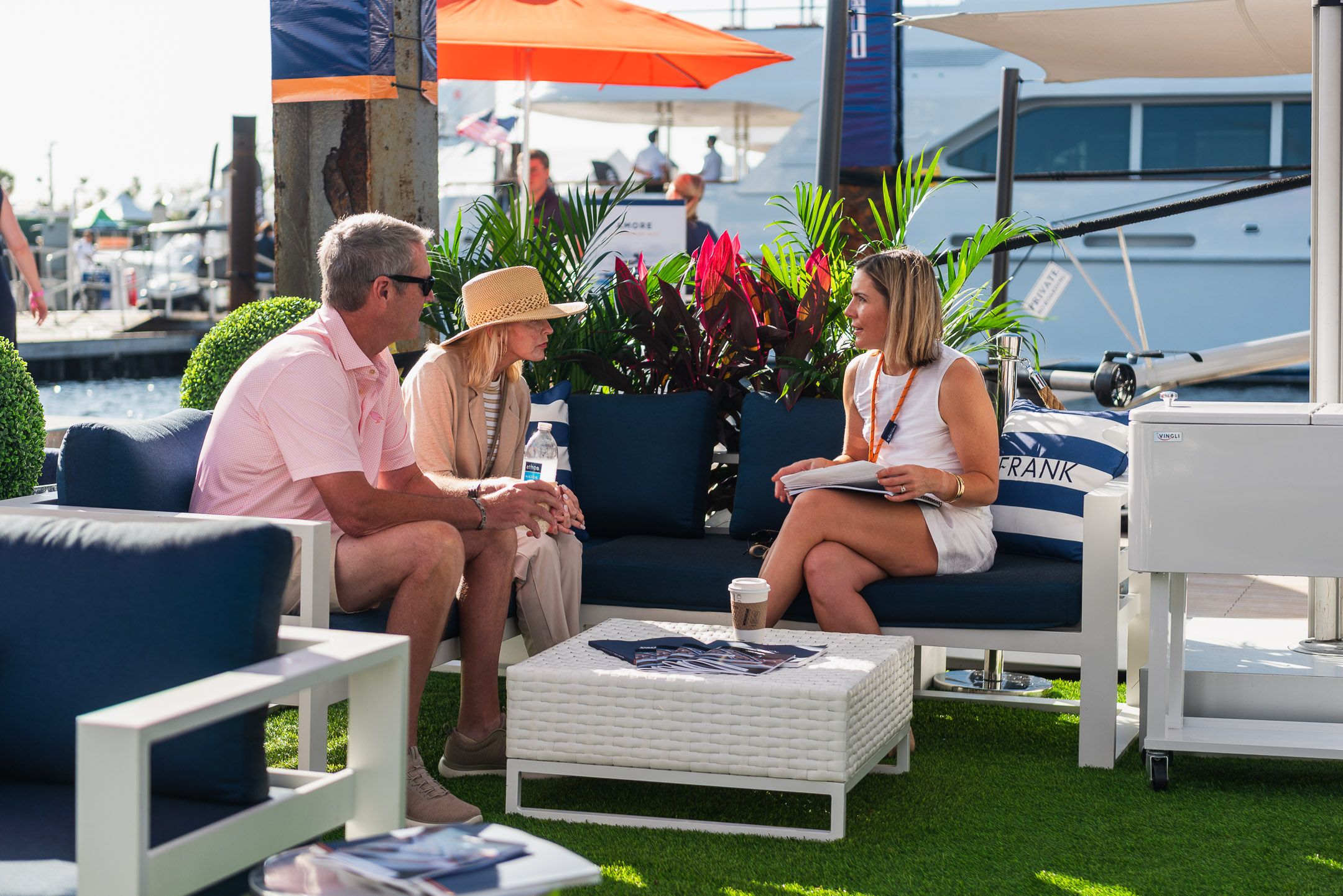 Three people converse on white outdoor patio furniture with blue cushions at a lively marina event.