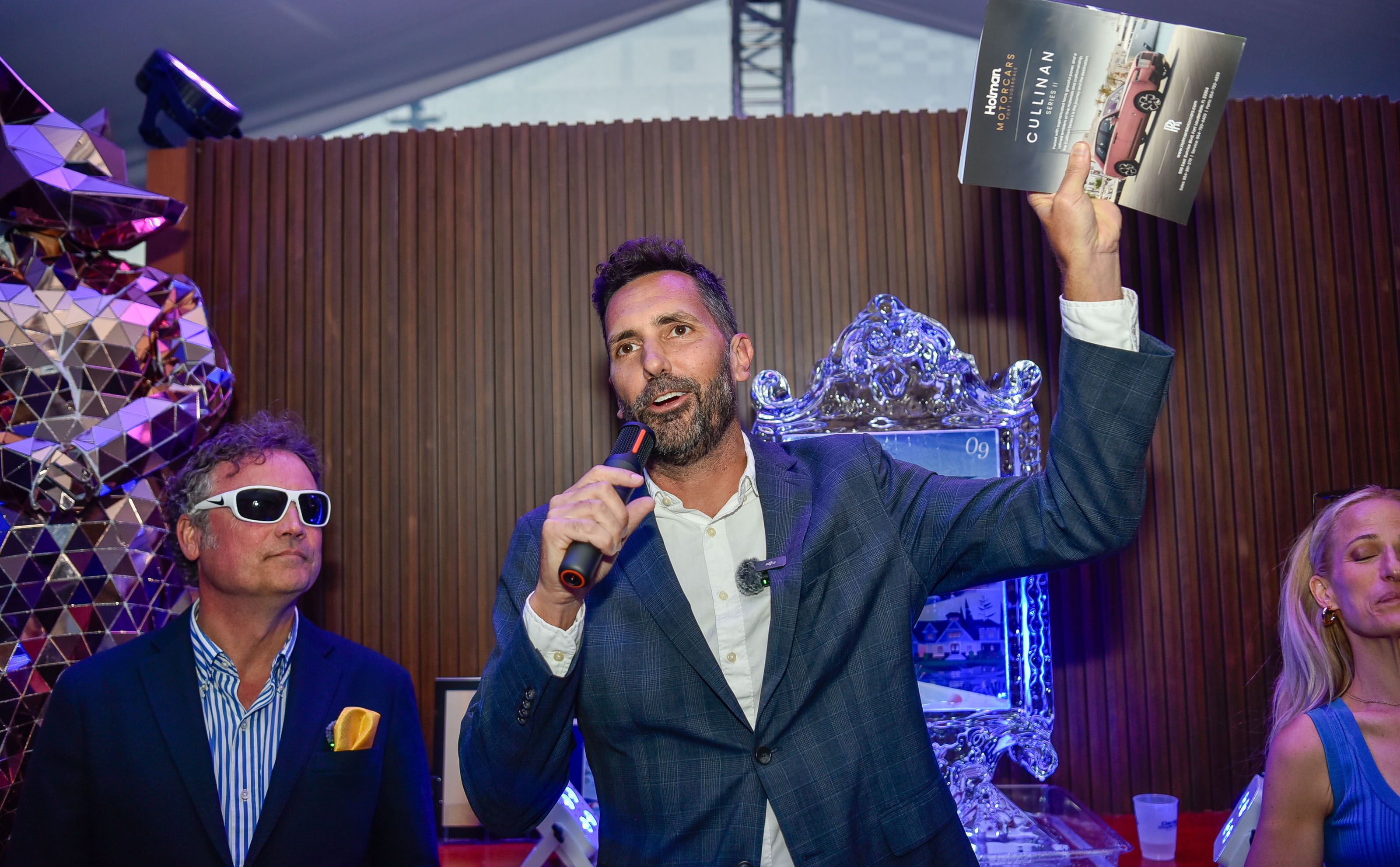 Man in suit speaks into microphone, holding a booklet at an event.