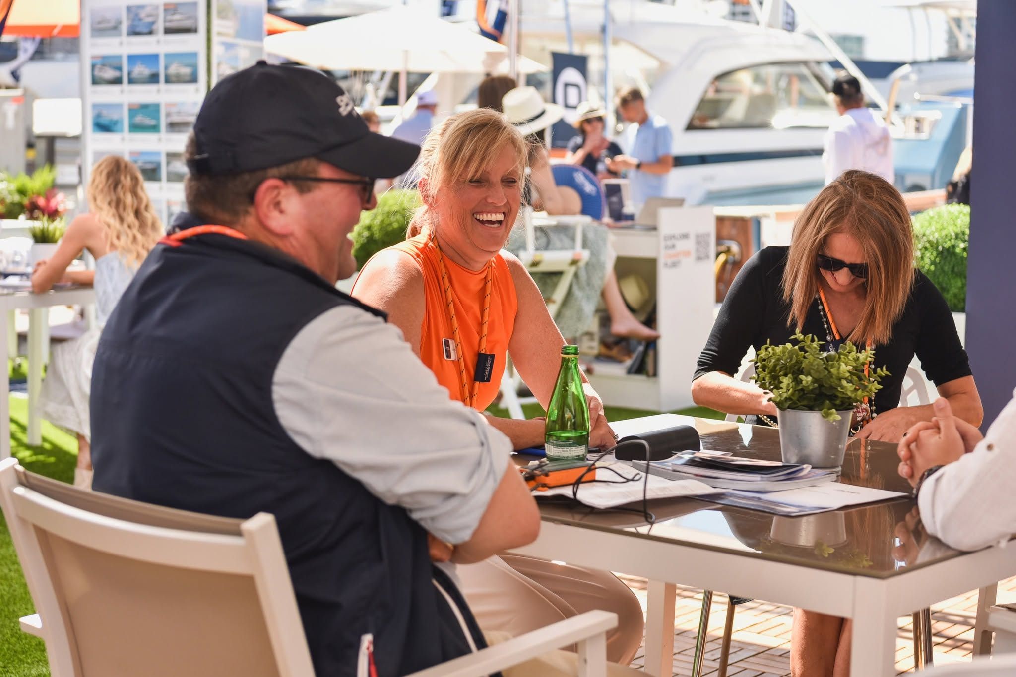 Three people laughing and socializing at an outdoor table with boats in the background.