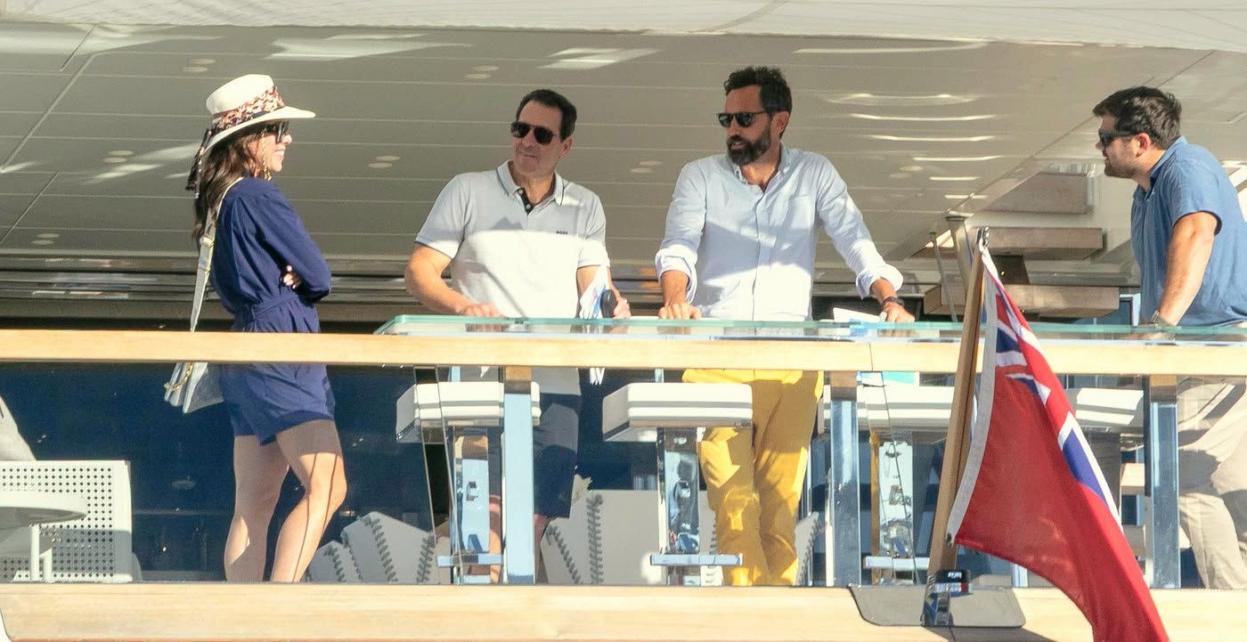 Four people on a yacht deck, two men behind a bar, British flag visible.