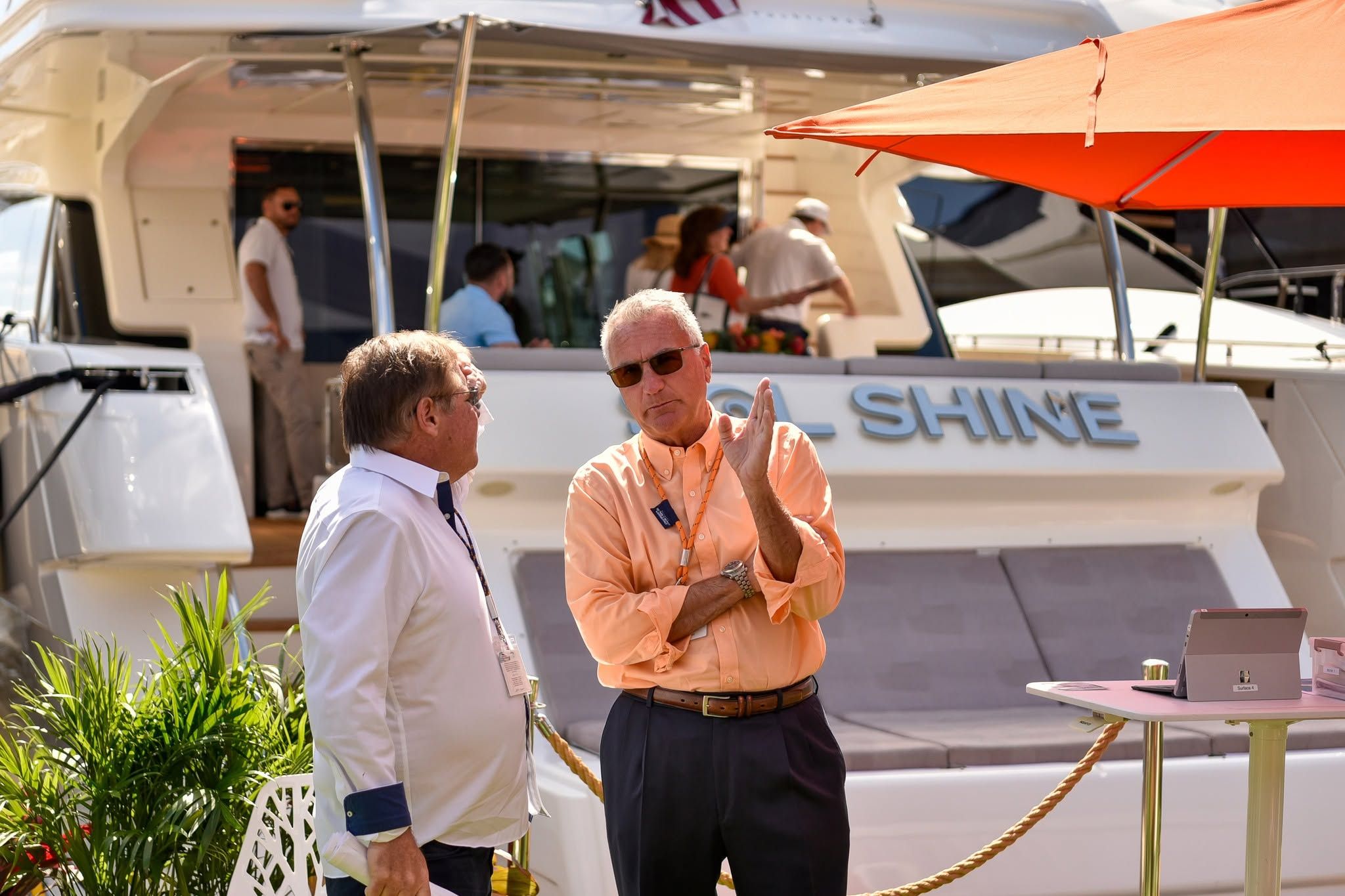 Two men converse in front of a large white yacht with people on board.