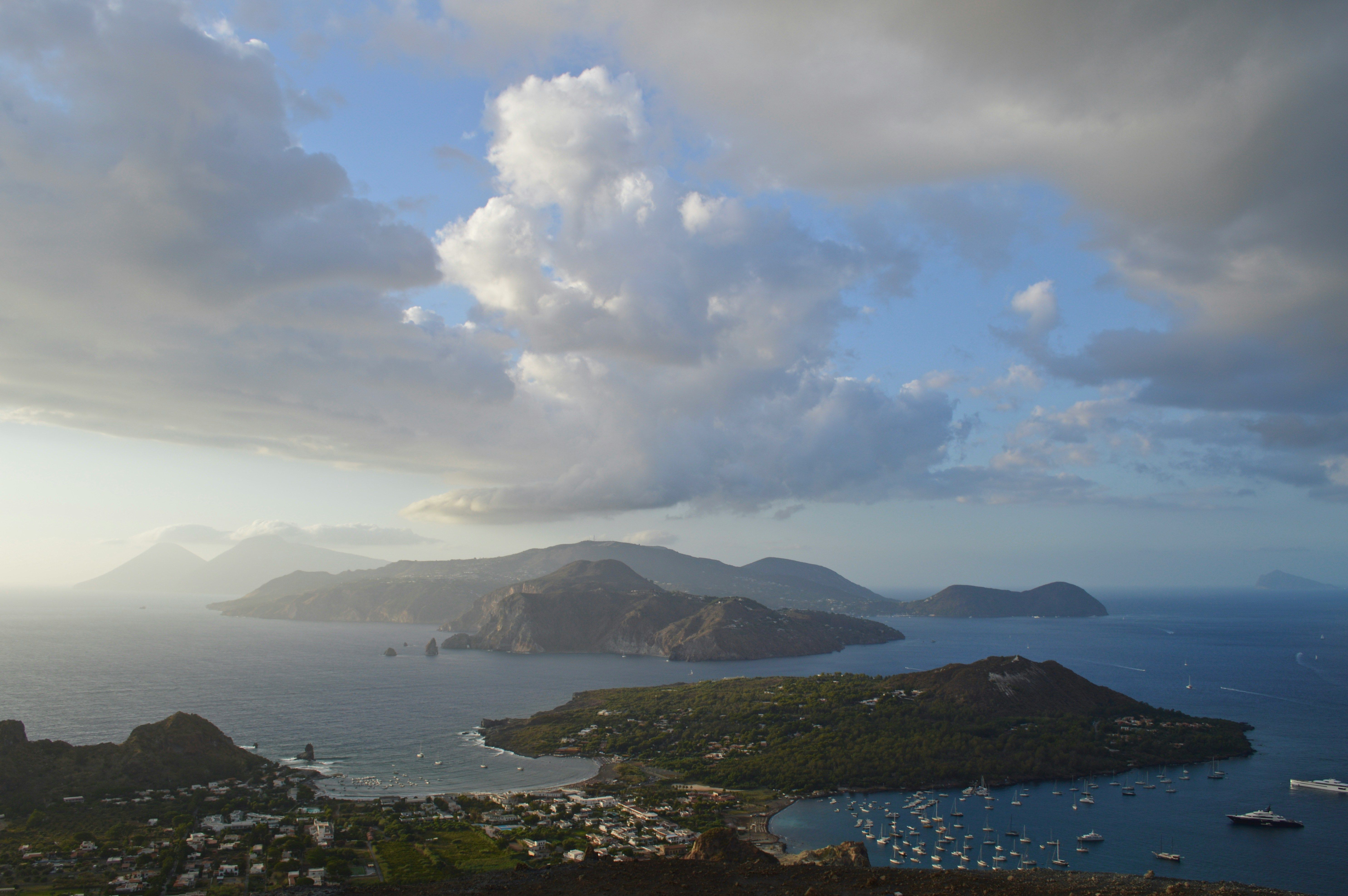Aerial view of volcanic islands, a town, and boats in a bay under a cloudy sky.