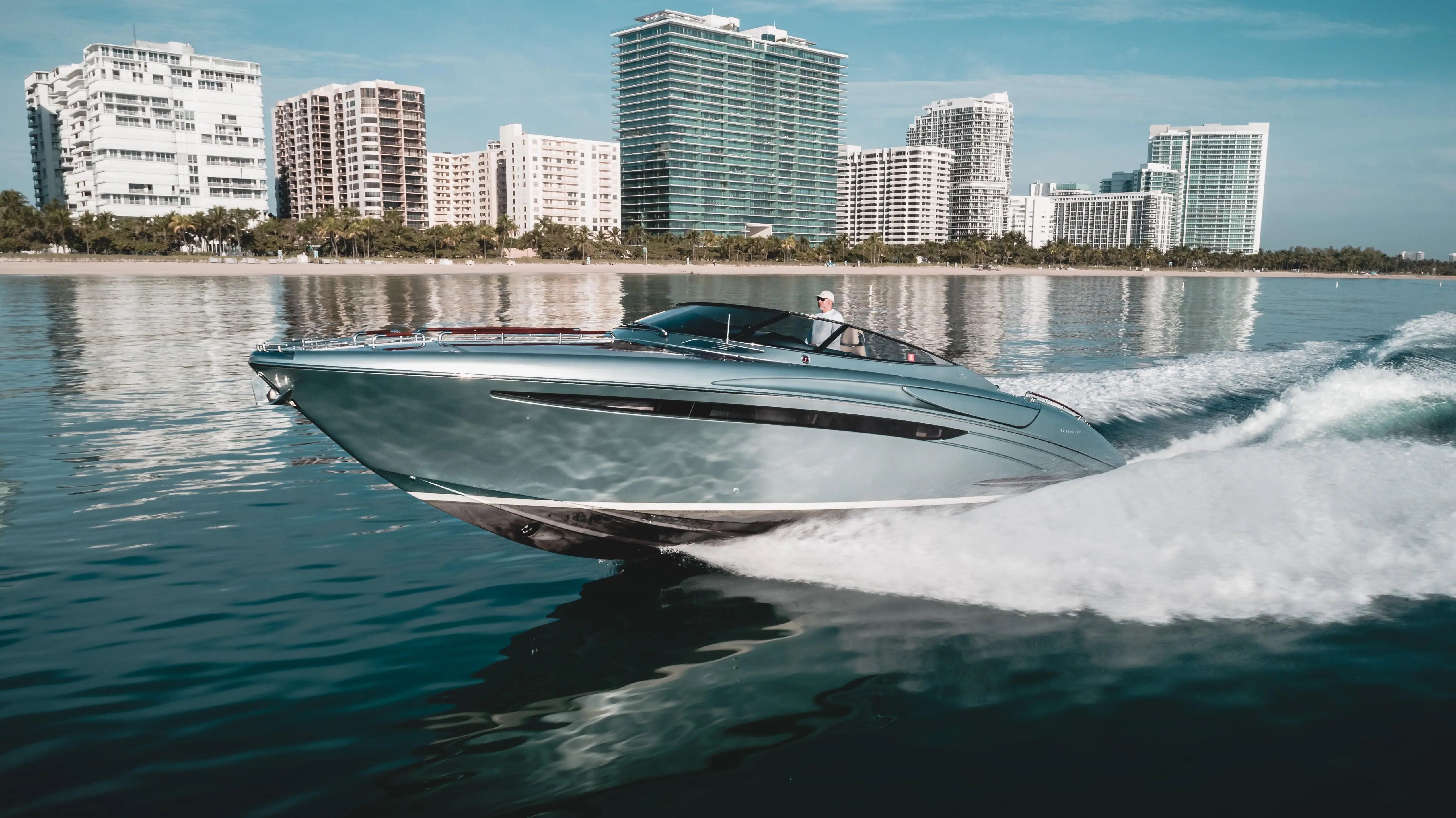 Sleek silver yacht speeds across blue water with a modern city skyline in the background.