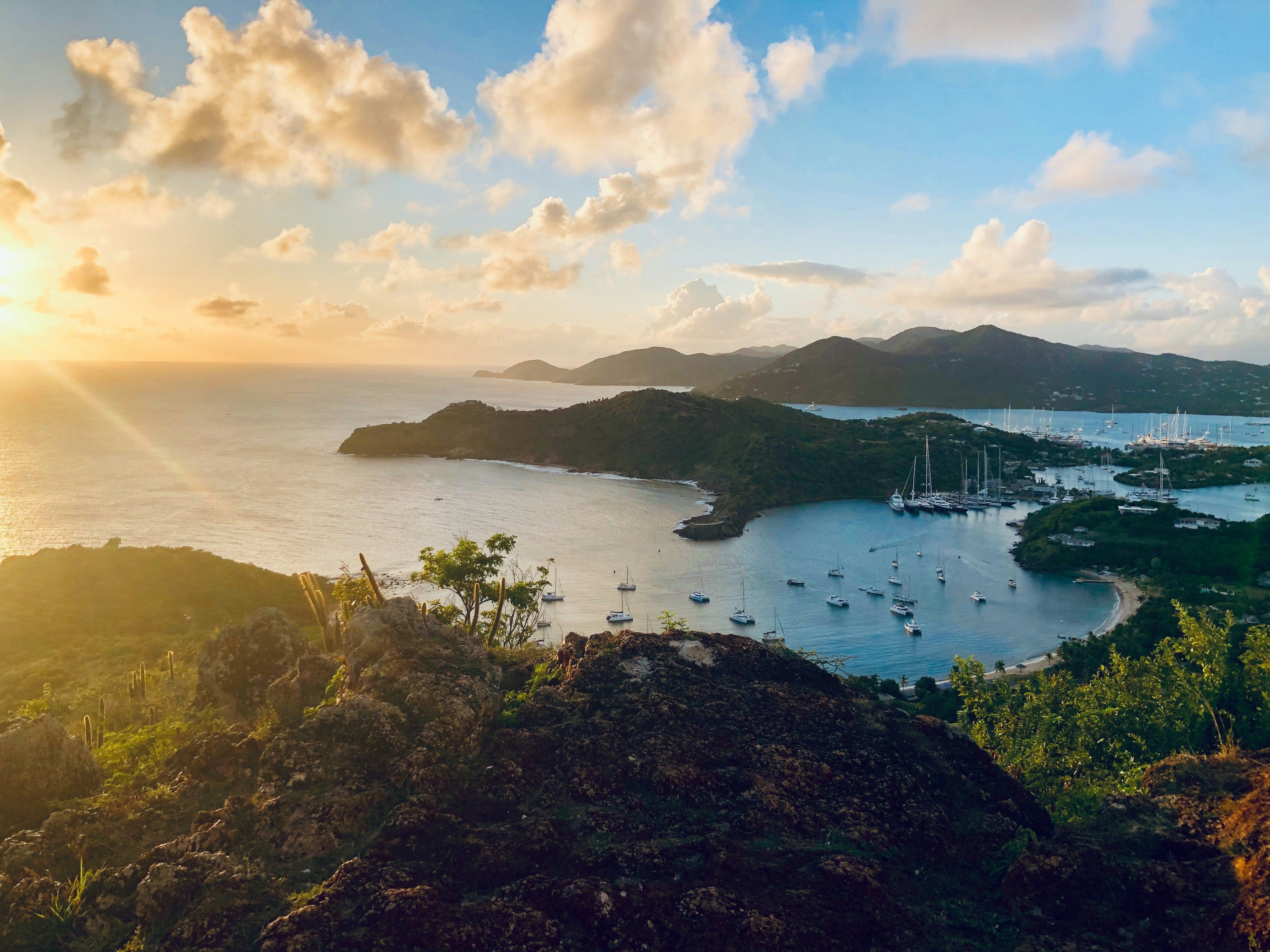 A picturesque bay filled with boats at sunset, surrounded by green hills under a cloudy sky.