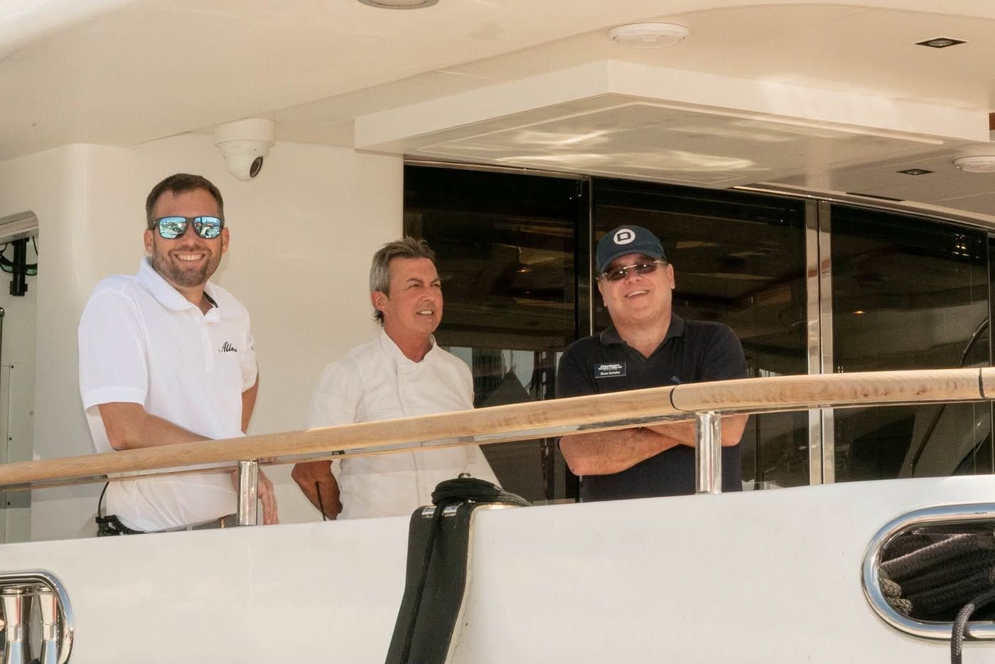 Three smiling men stand on a yacht deck, two wearing sunglasses, looking forward.
