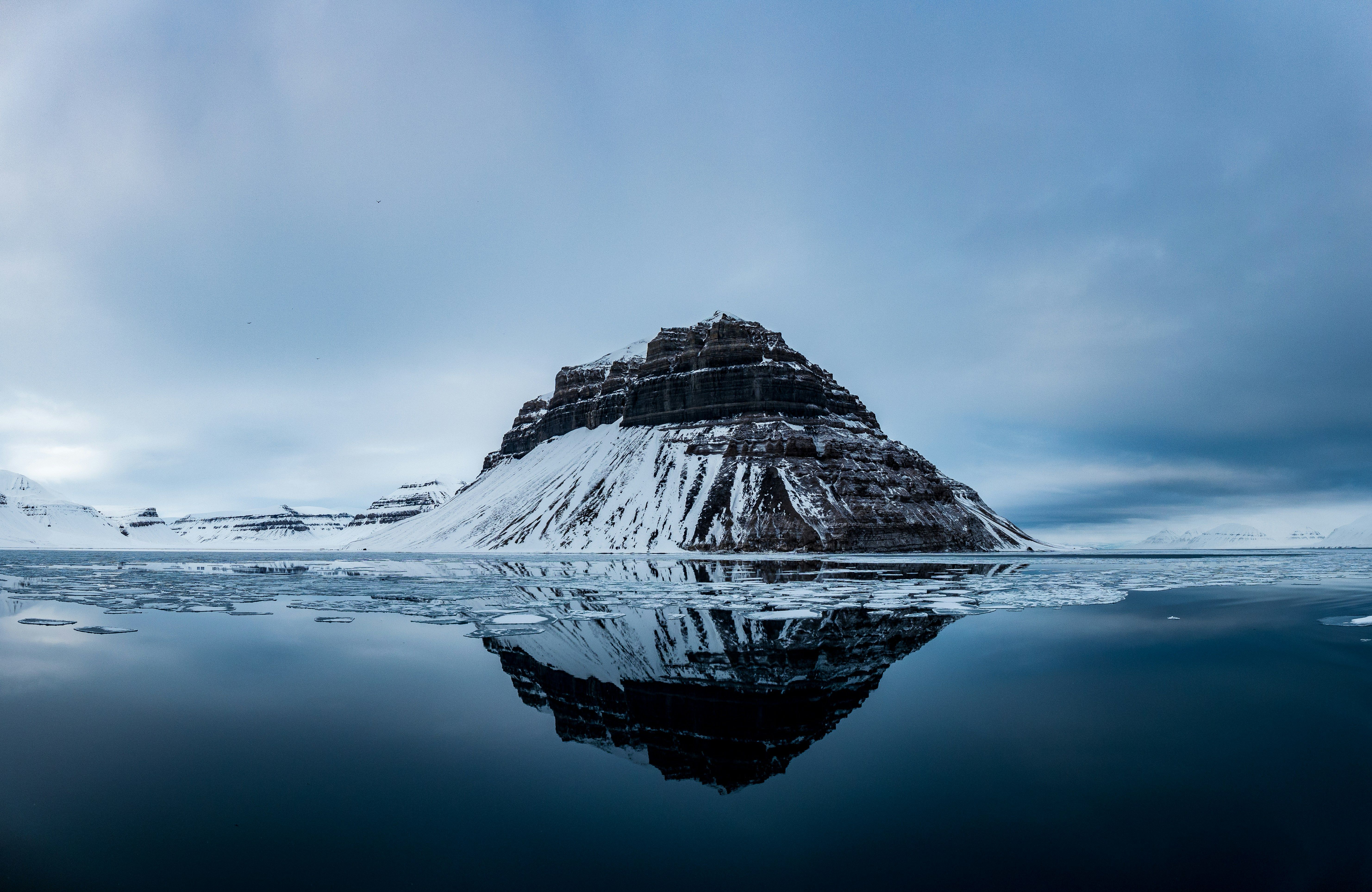 Snow-capped rocky mountain reflected in still, dark water under a cloudy sky.