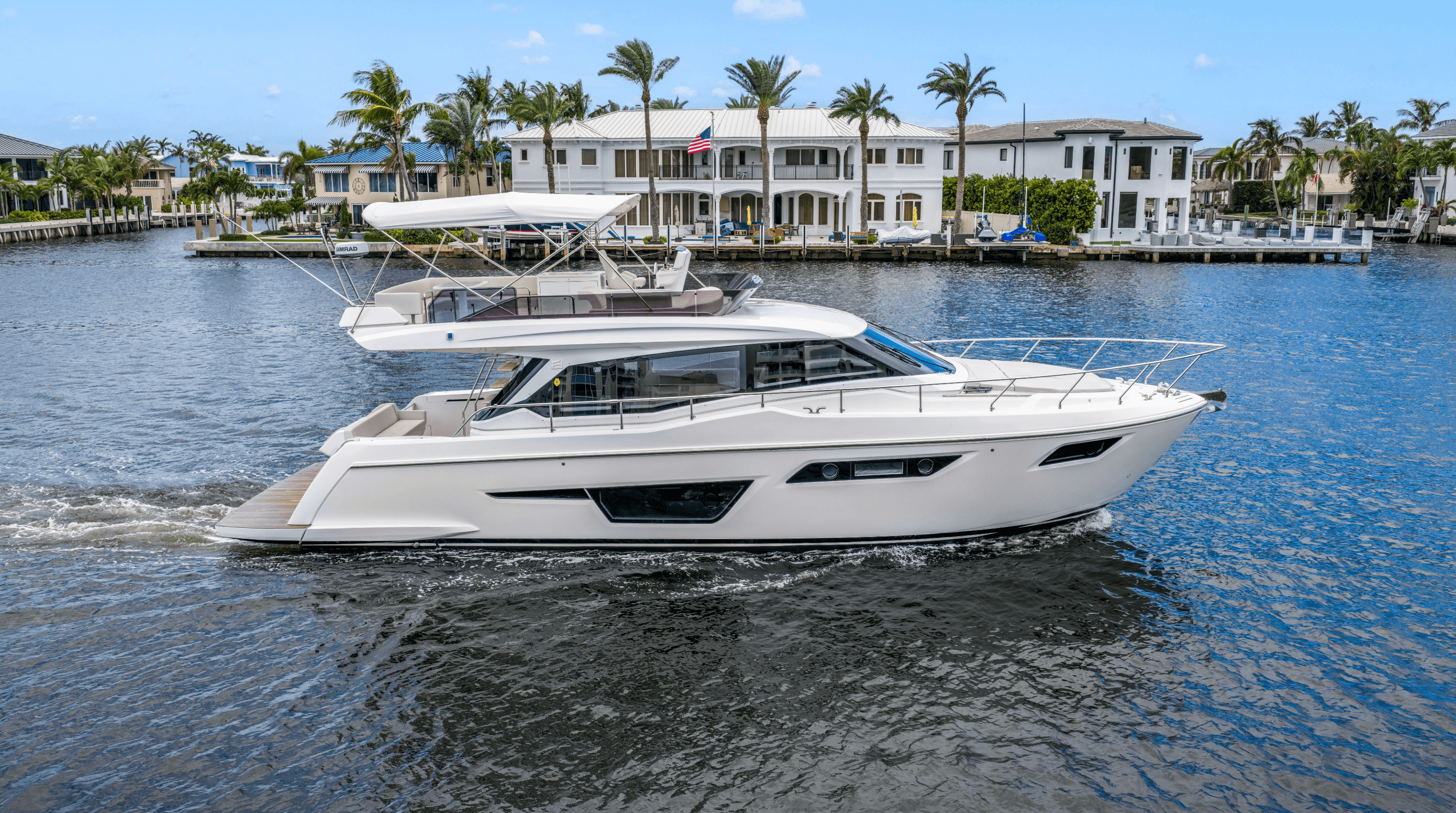 A sleek white luxury yacht cruises on a blue waterway past waterfront homes and palm trees.