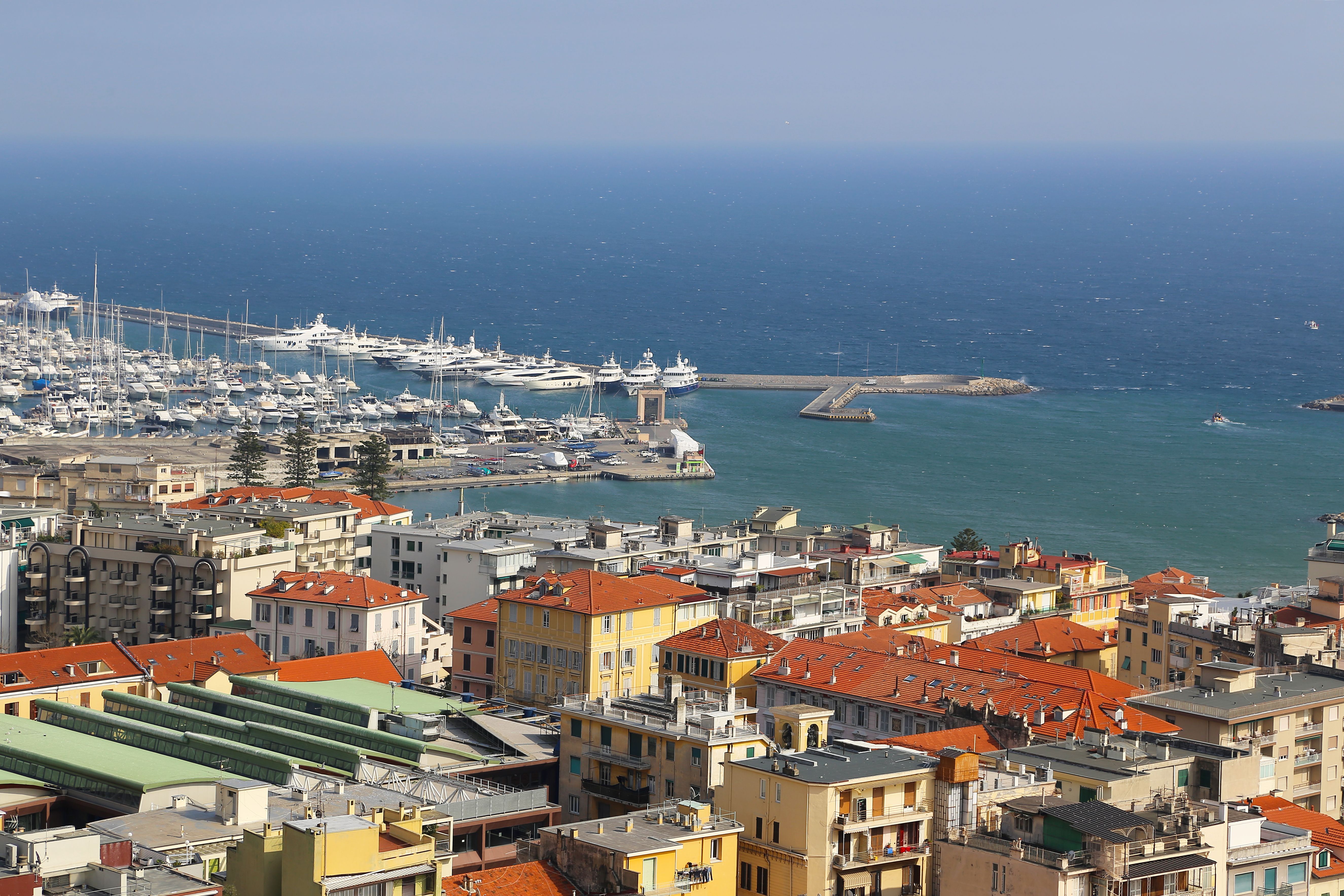 A vibrant coastal city with red-roofed buildings overlooking a busy marina and blue sea.