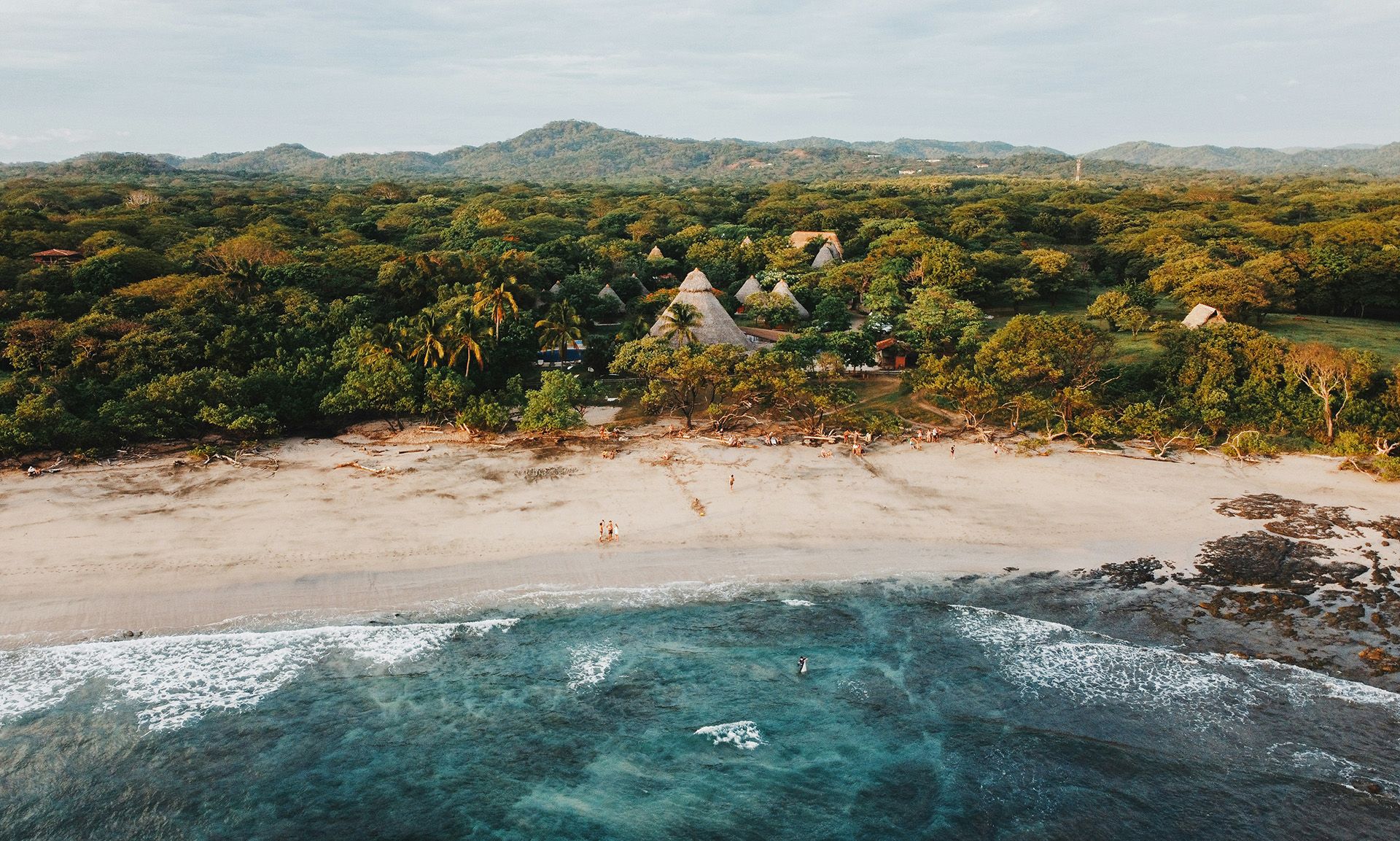 Aerial view: tropical beach with turquoise ocean, dense forest, and unique buildings.