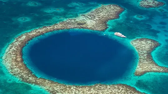 Aerial view of a large, circular dark blue sinkhole surrounded by coral reefs and a boat.