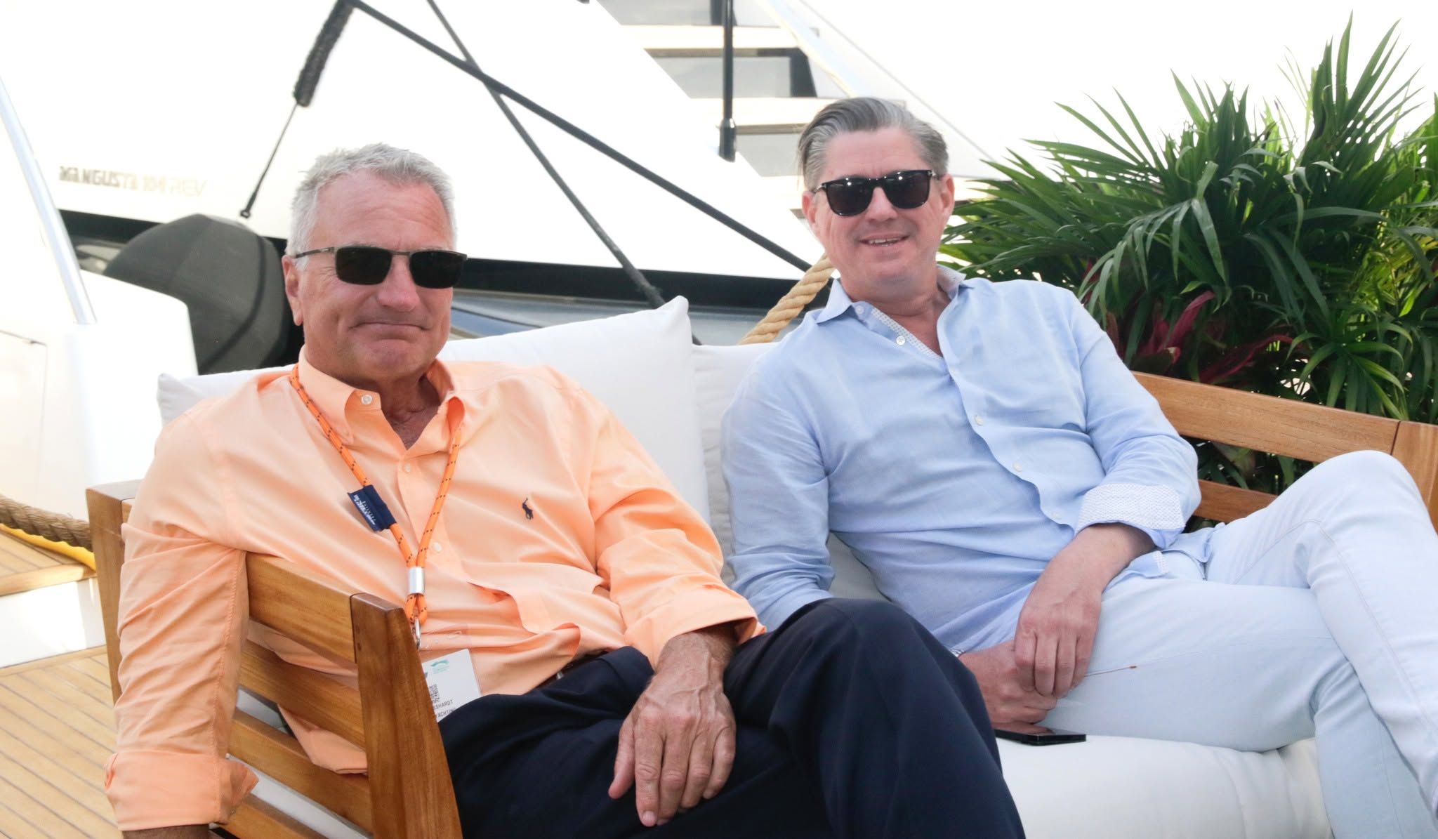 Two men in sunglasses relax outdoors, one in a peach shirt, the other in a blue shirt.