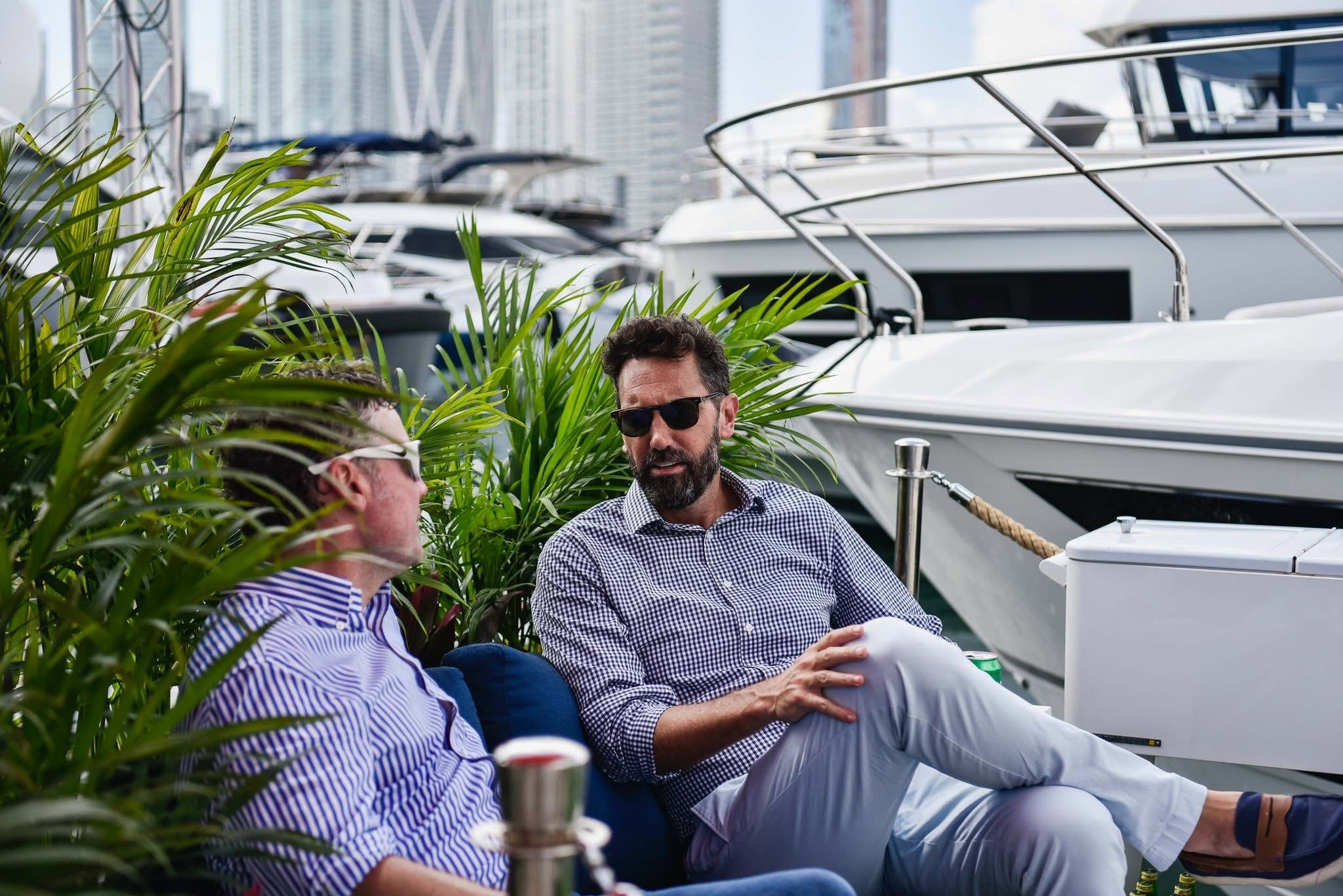 Two men converse outdoors on a sunny marina deck with large boats nearby.