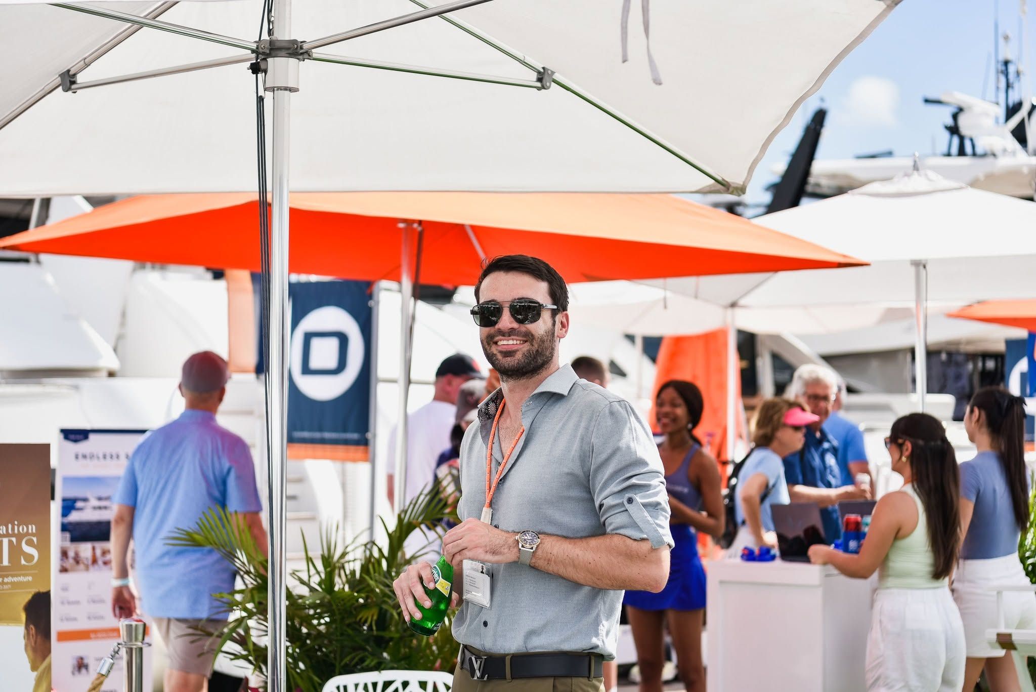 A man in sunglasses and a grey shirt smiles, holding a drink at an outdoor event.