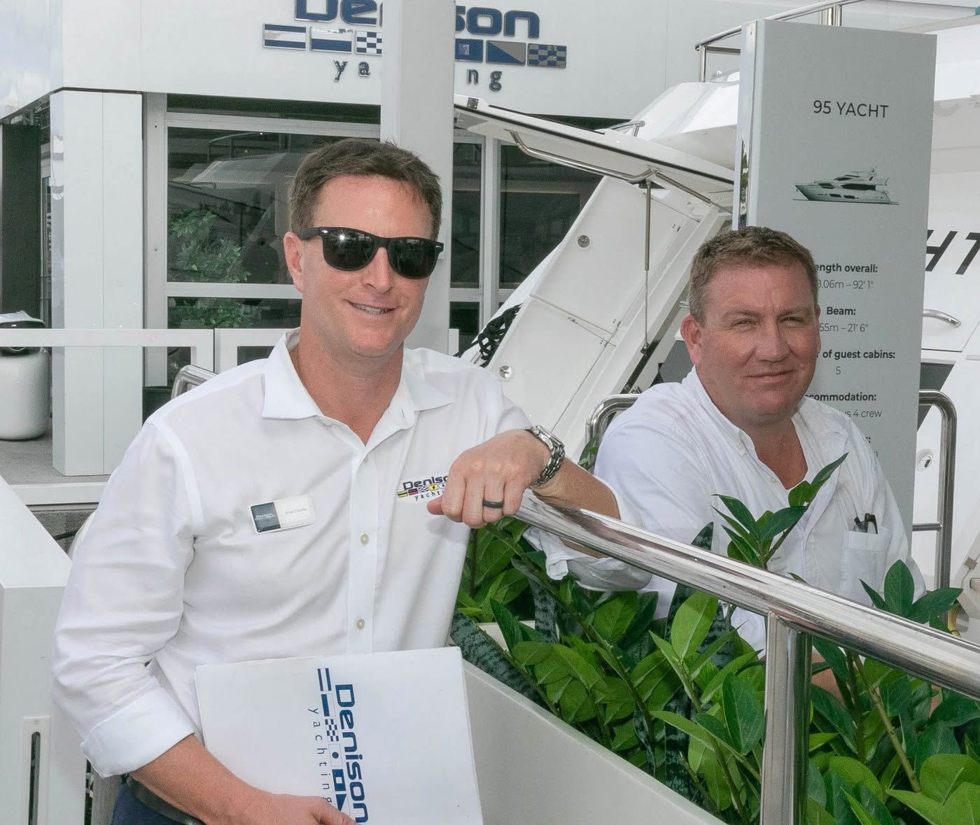 Two men in white shirts and sunglasses smile while standing near a Denison yacht display.