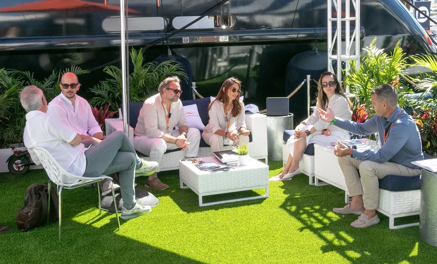 Six people, four men and two women, are sitting on a green outdoor area, talking.