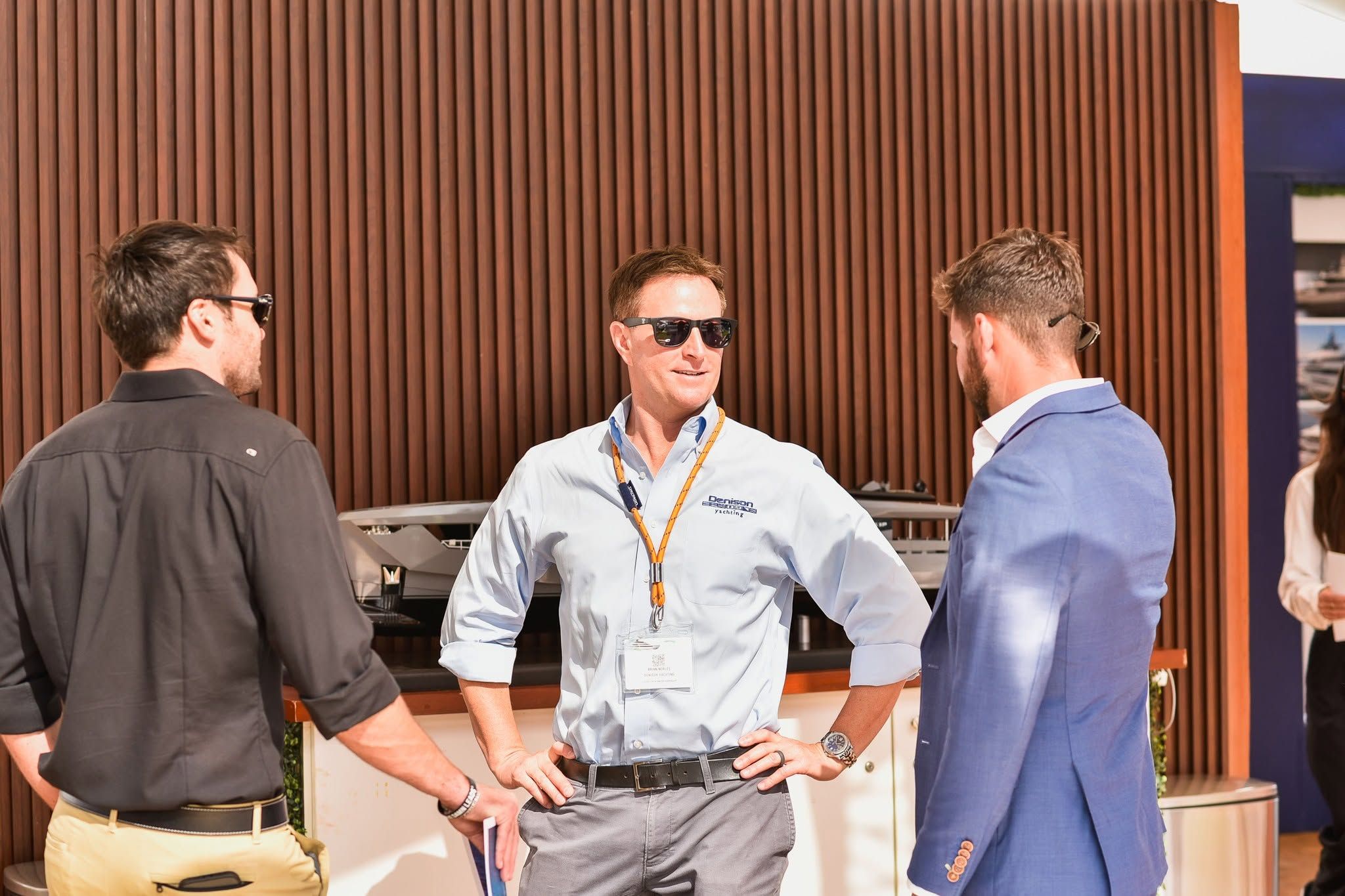 Three men in sunglasses converse in a professional setting with a wooden panel backdrop.