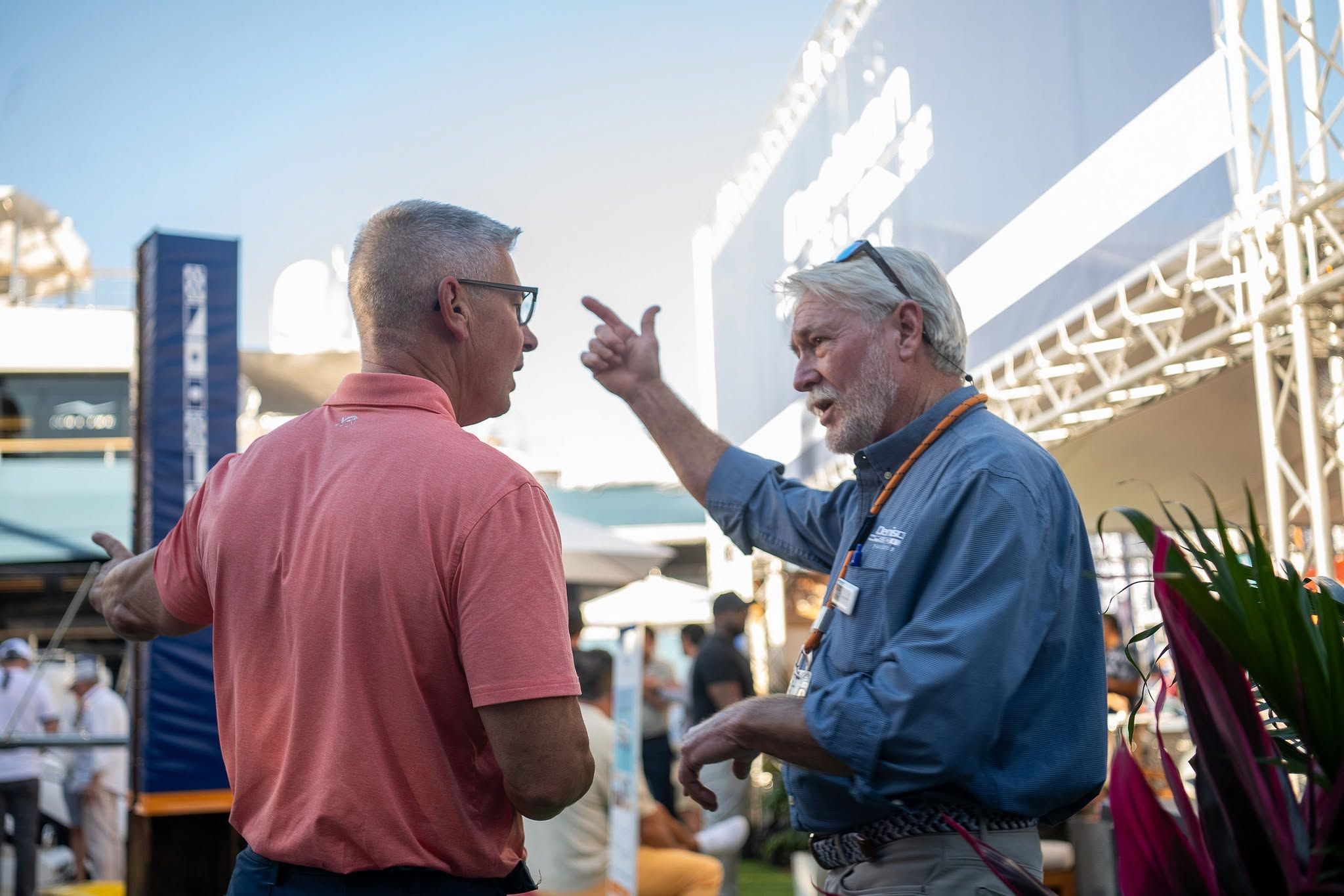 Two men, one in a blue shirt pointing, engage in a conversation at an outdoor event.