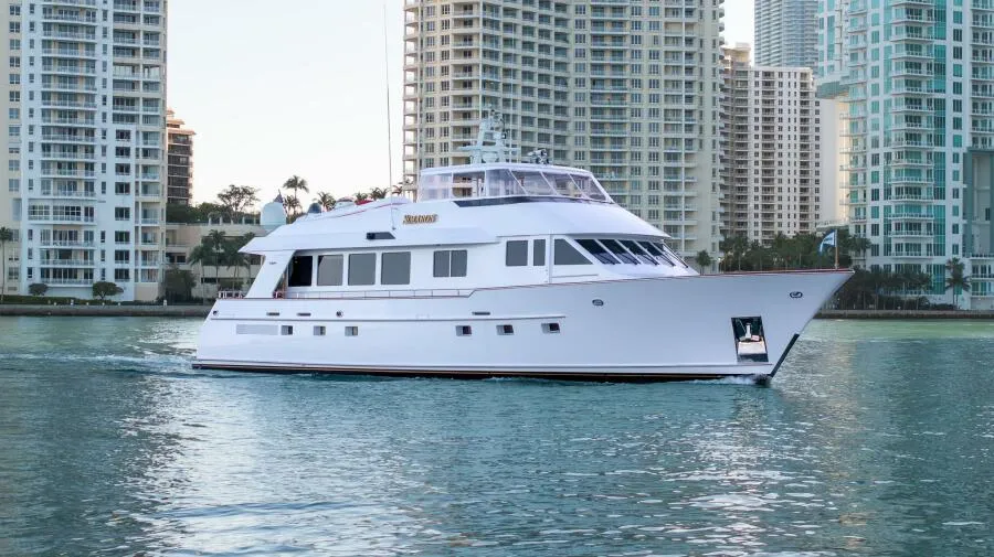 A sleek white yacht sails on calm water with tall city buildings in the background.
