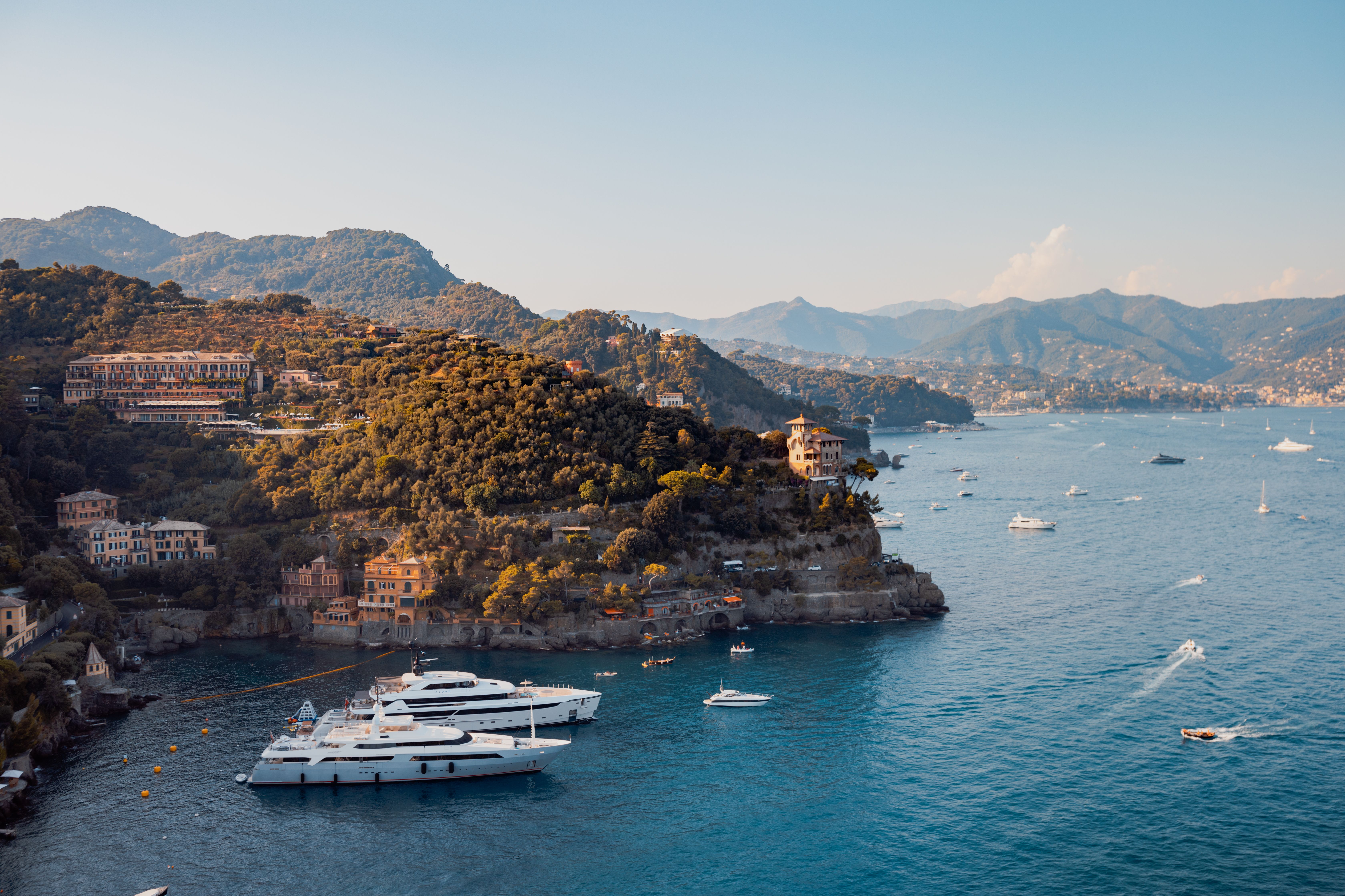 Scenic coastal bay with luxury yachts, green hills, and buildings under a clear sky.