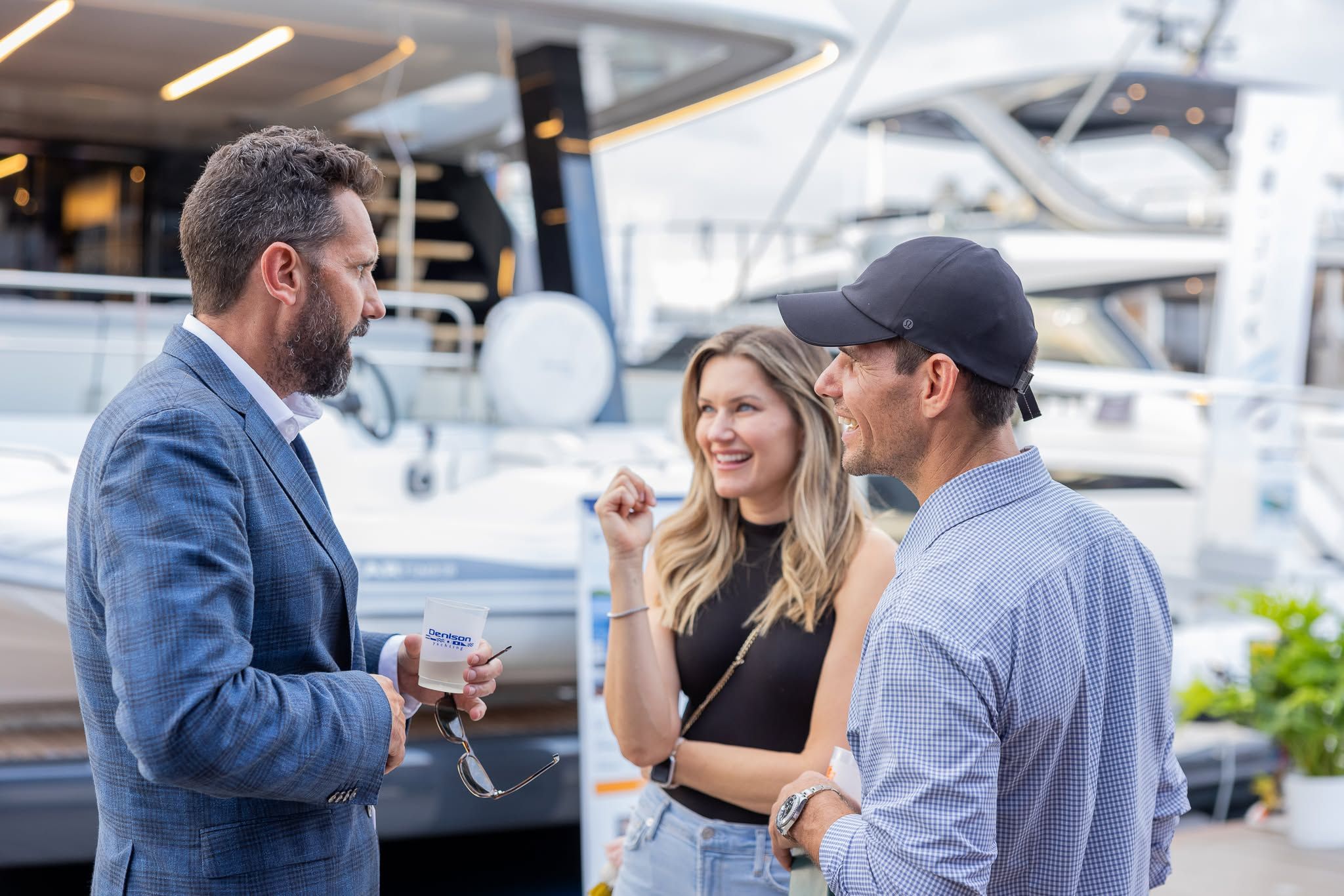 Three people conversing at a yacht show, smiling with luxury boats behind them.