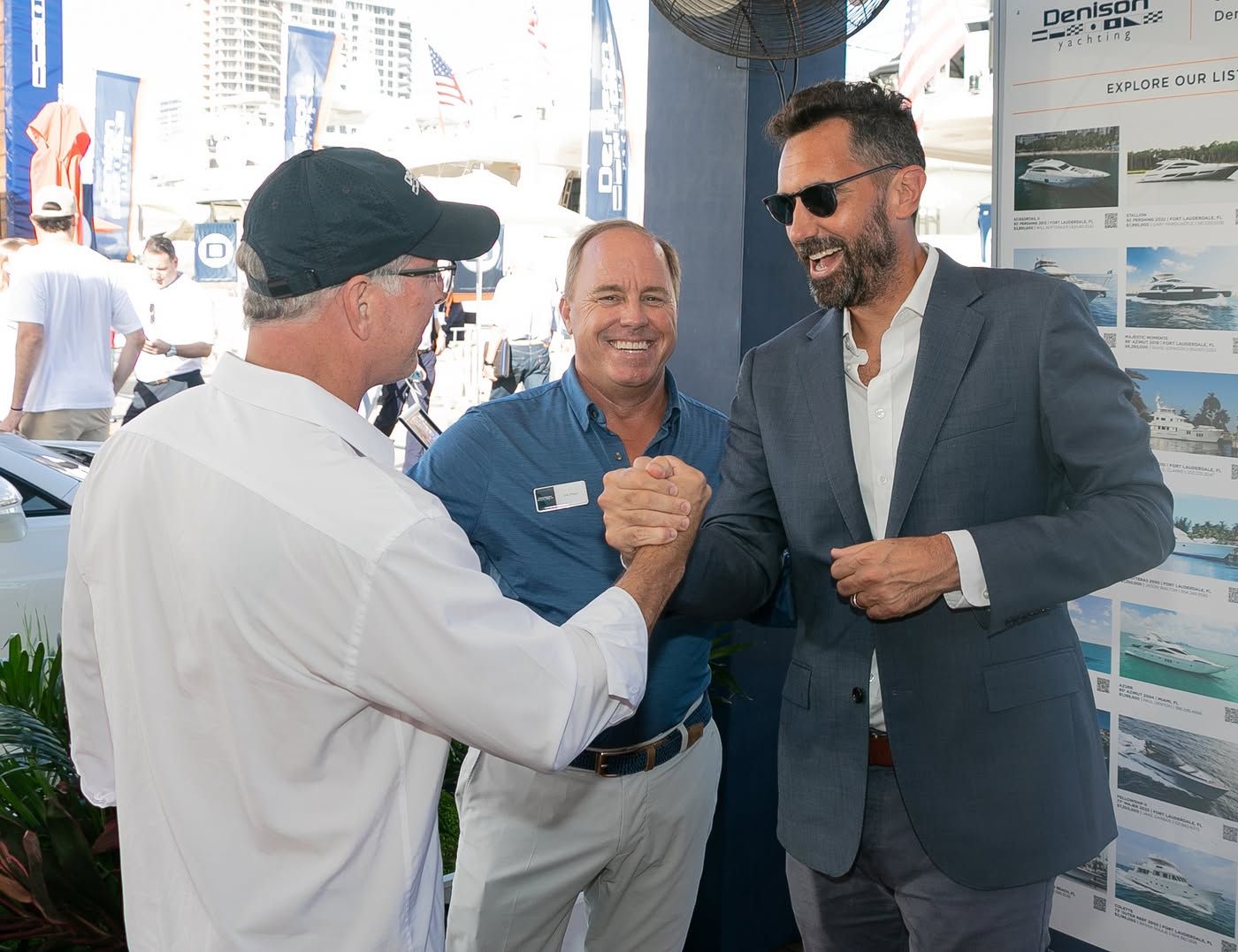 Three men smile and shake hands at an outdoor event with boat displays in the background.