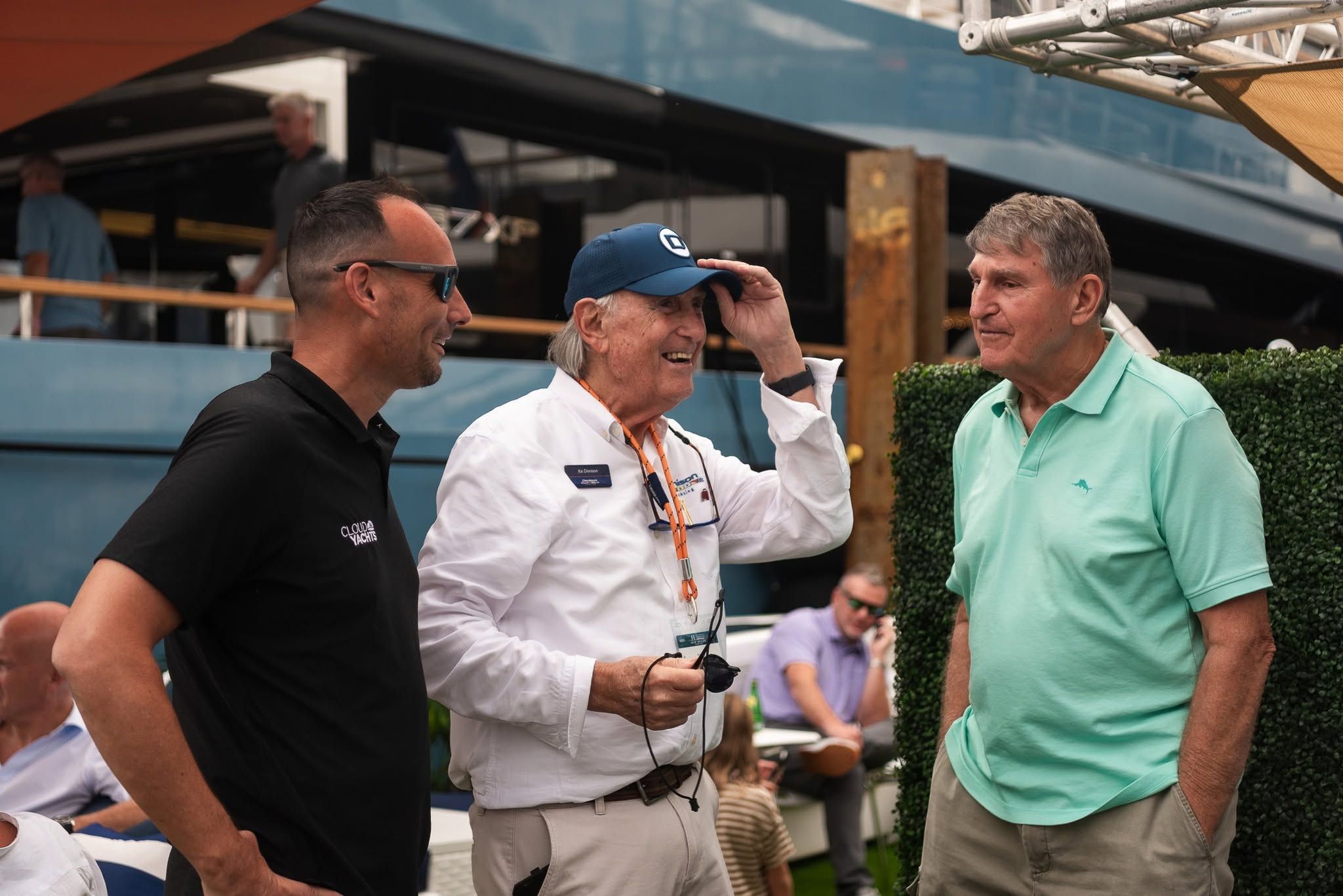 Three men smile and converse outdoors, one adjusting his blue cap at a social event.