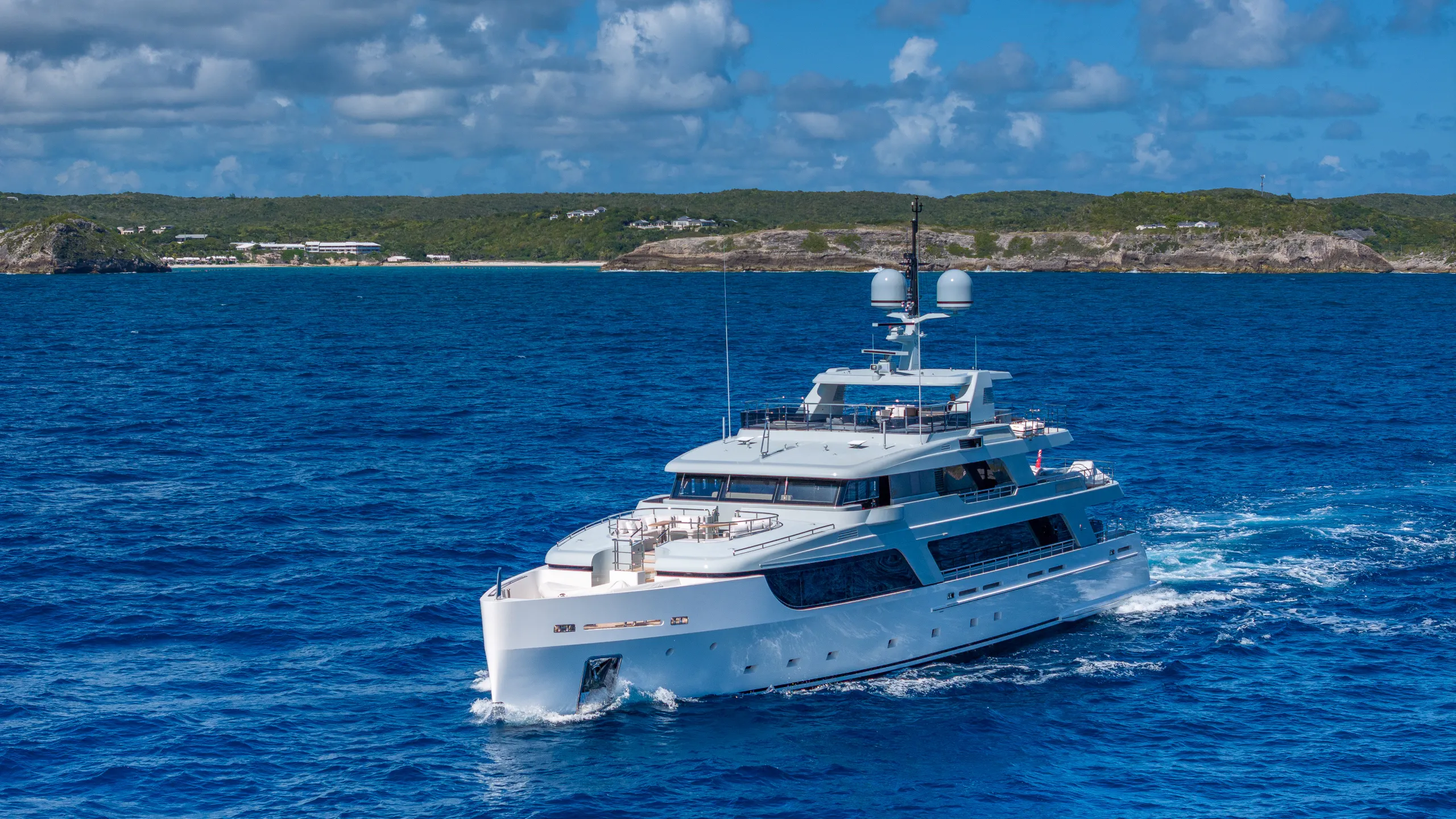Large white luxury yacht sails across deep blue water near a green tropical island.
