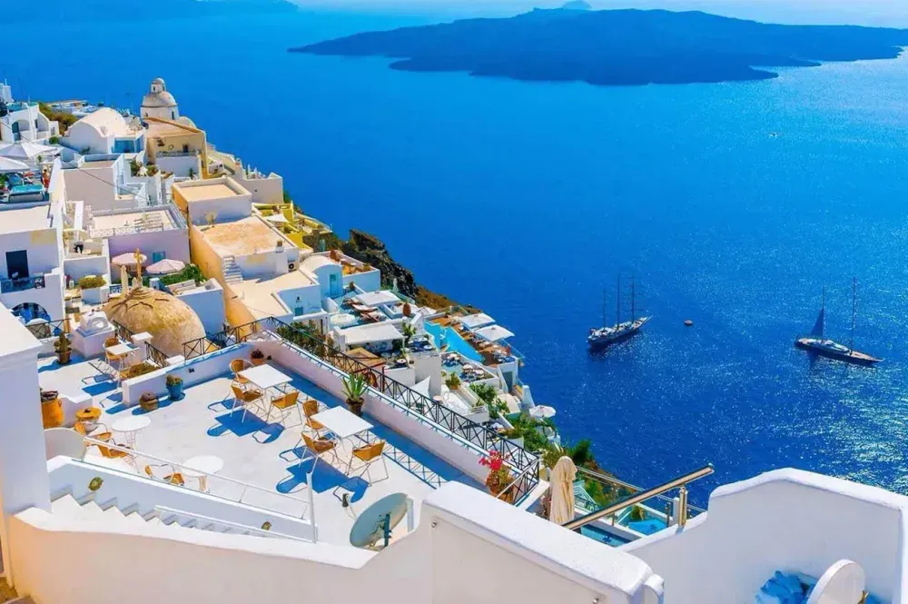Whitewashed buildings on a cliff overlook the deep blue Aegean Sea with boats.