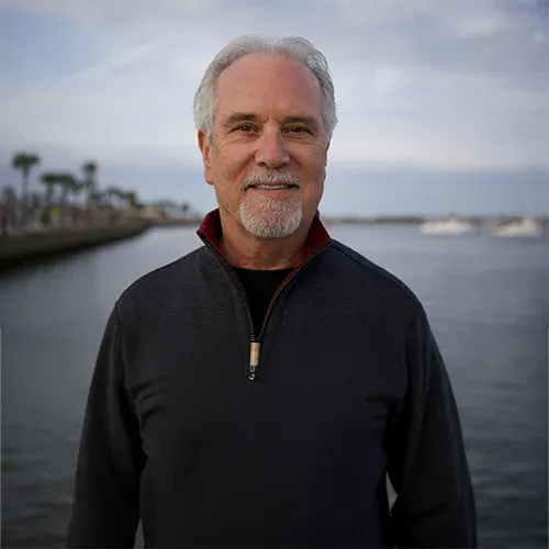 A smiling man with gray hair and a goatee stands by the water.