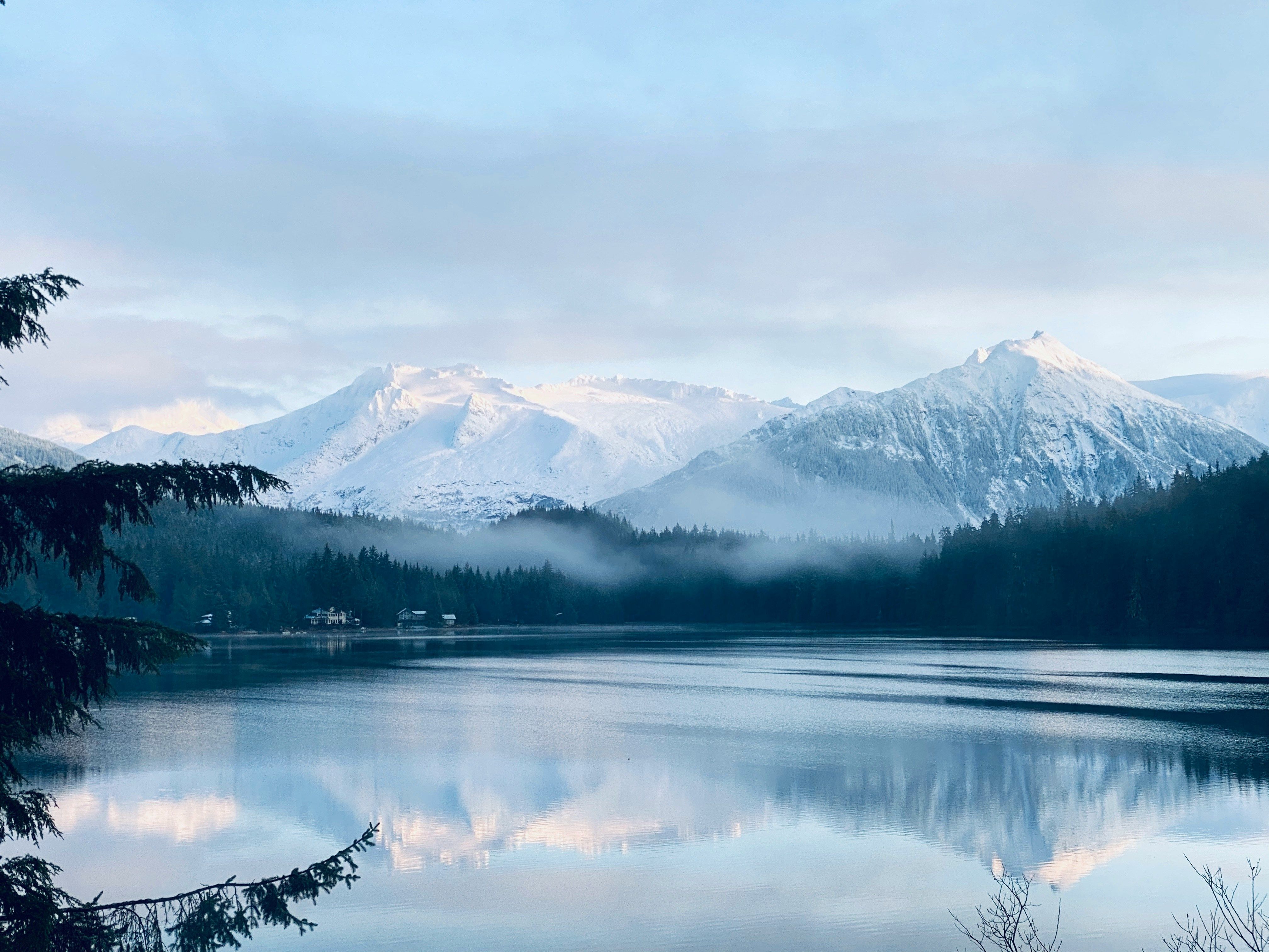 Serene lake reflecting snow-capped mountains under a cloudy sky, framed by dark trees.