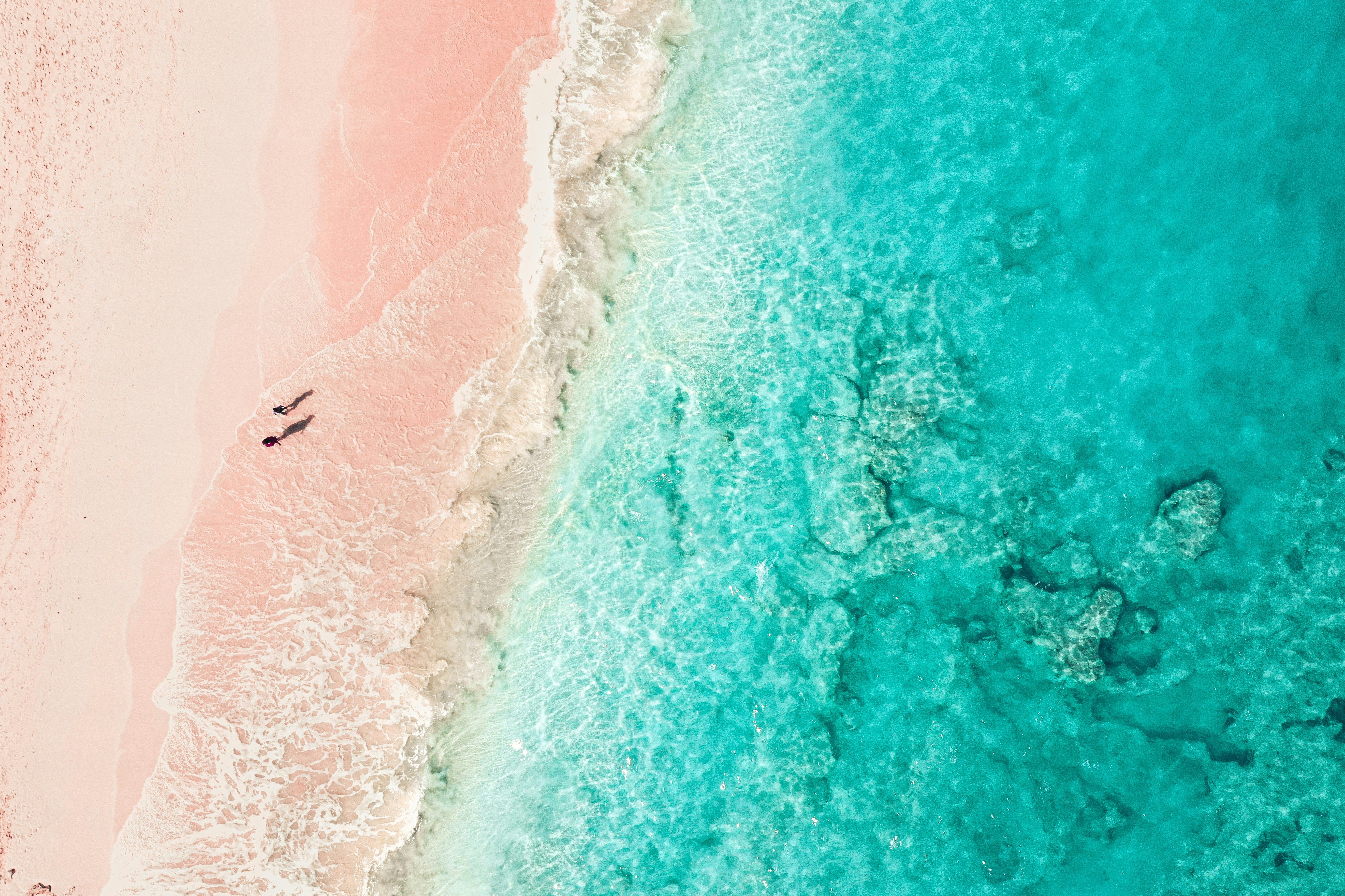 Aerial view of a pink sand beach meeting clear turquoise ocean with two tiny figures.