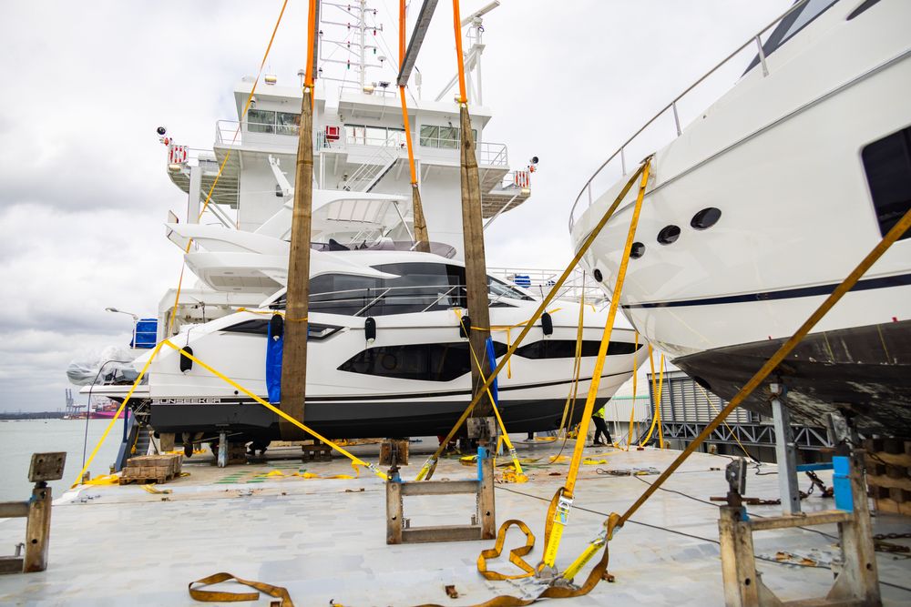 White yacht carefully lifted by straps onto a large transport ship for delivery.