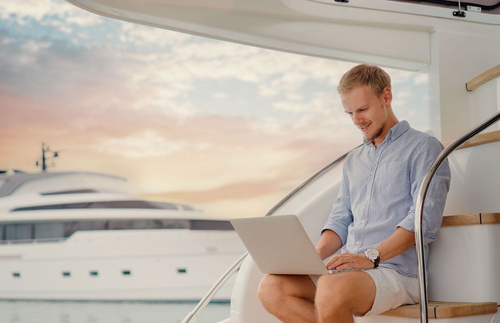 Man works on a laptop on a luxury yacht with a sunset backdrop.