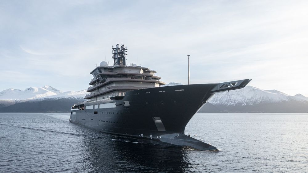 A sleek, dark superyacht cruises through calm waters with snowy mountains in the background.