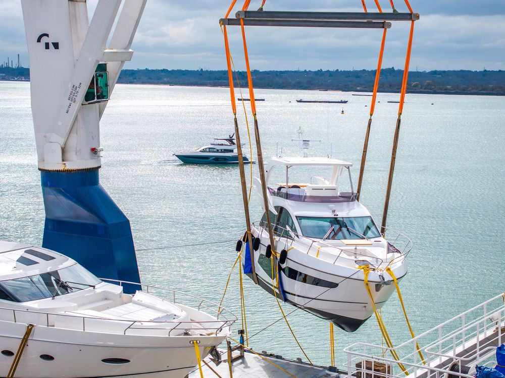 A white yacht is lifted by a crane from a transport ship over the water.