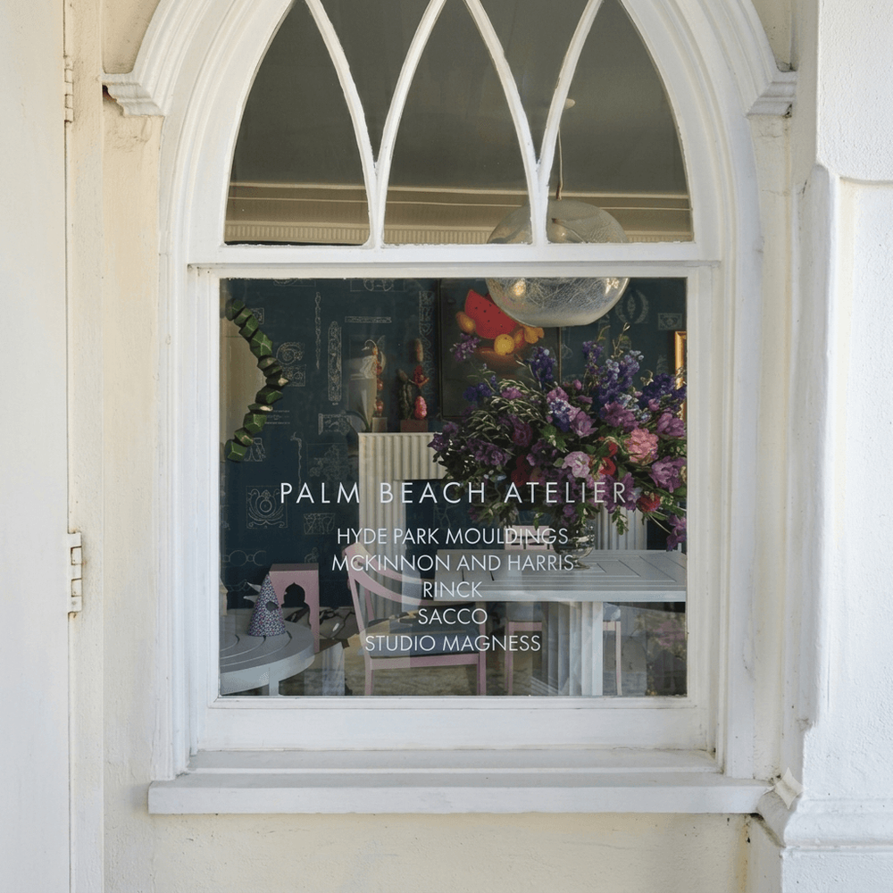 White arched storefront window of Palm Beach Atelier showcasing interior decor and flowers.