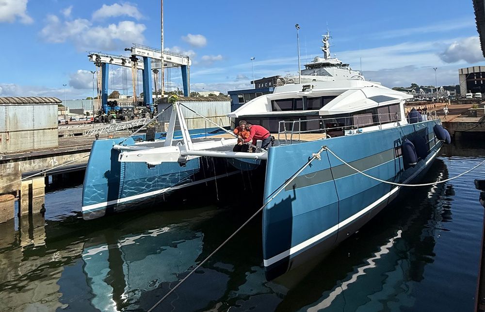 Large blue and white catamaran in a shipyard dry dock with workers on deck.